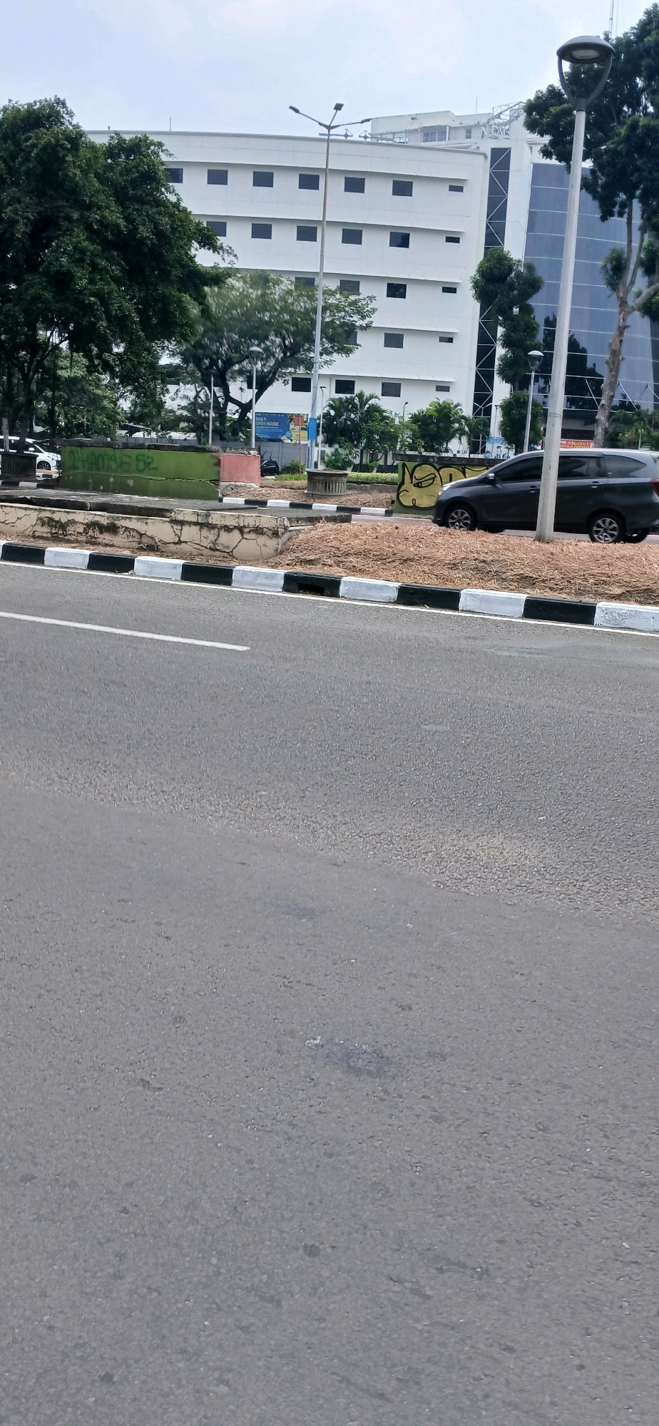 The image shows a view of a street in Central Jakarta, Indonesia. In the foreground, there is a paved road with a white line marking the lane. The median of the road is decorated with alternating black and white painted concrete curbs. Beyond the median, there is a grass area, and further back, a dark-colored car is parked. Behind the car, there is a building with a modern architectural design, characterized by a white facade and numerous dark windows. Trees and a lamppost are also visible in the scene, contributing to the urban landscape. The sky is overcast.