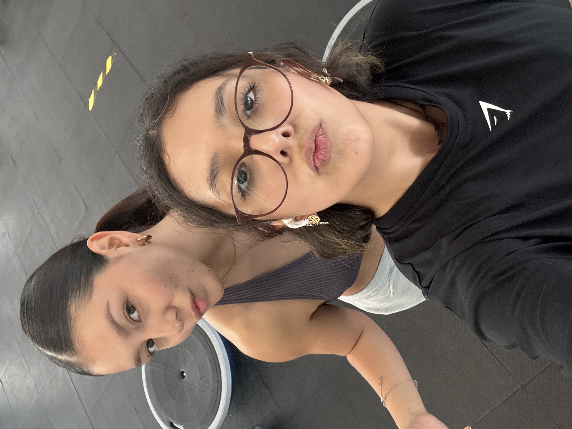 Two young women are captured in a close-up selfie within an indoor setting, likely a gym or fitness facility, given the dark rubber mat flooring and visible gym equipment. The image is taken from a slightly upward angle, with both subjects looking towards the camera.
In the foreground, a woman with dark hair, styled down, wears round, brown-rimmed glasses. She has a playful "duck face" expression. She is wearing a black t-shirt with a distinctive white logo on the chest, an upward-pointing triangular shape often associated with the brand Gymshark. She also has elaborate white and gold earrings and a delicate bracelet on her wrist.
Partially behind her, slightly to the left, is another young woman with dark hair pulled back into a ponytail. She is wearing a dark, ribbed sports bra or top and light-colored bottoms (possibly shorts). She looks towards the camera with a subtle pout and has a gold hoop earring visible.
The background features a dark grey or black floor with some bright yellow diagonal striped markings. A circular piece of gym equipment, possibly a weight plate or part of a machine, is partially visible behind the second woman. The overall mood of the image is casual and playful, suggesting a break or pause during a workout. The location context provided is San Andrés Cholula, Mexico.