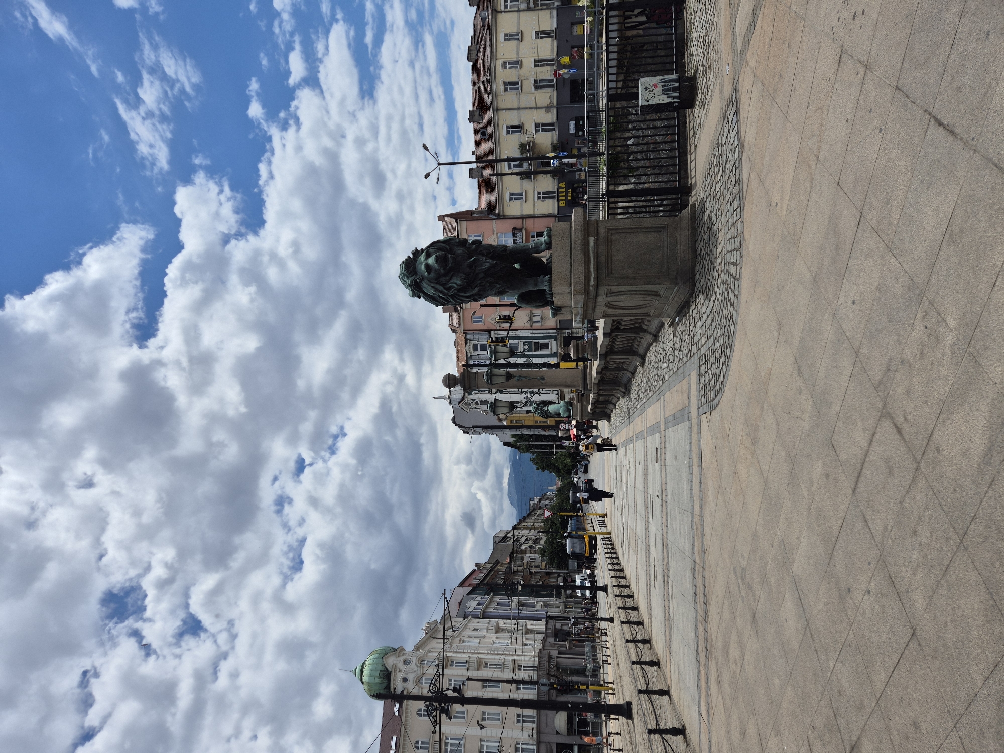 The image shows the Lion's Bridge (Lviv Most) in Sofia, Bulgaria, on a cloudy day. A wide pedestrian walkway lines the street, bordered by black metal bollards and chains. On the right side of the walkway, a large bronze lion statue sits atop a stone pedestal, overlooking the street. Beyond the statue, a metal fence separates the walkway from a lower area, possibly a canal or riverbed.
The street itself is paved with cobblestones and accommodates a yellow tram or bus. Several cars and pedestrians are visible on the street and walkway. Buildings with traditional European architecture, characterized by multiple stories, balconies, and pitched roofs, line both sides of the street. Some buildings feature ornate facades and decorative elements, such as a green dome on the left side. In the distance, mountains are visible under the cloud-filled sky. The overall scene depicts a busy urban environment with historical architectural features.