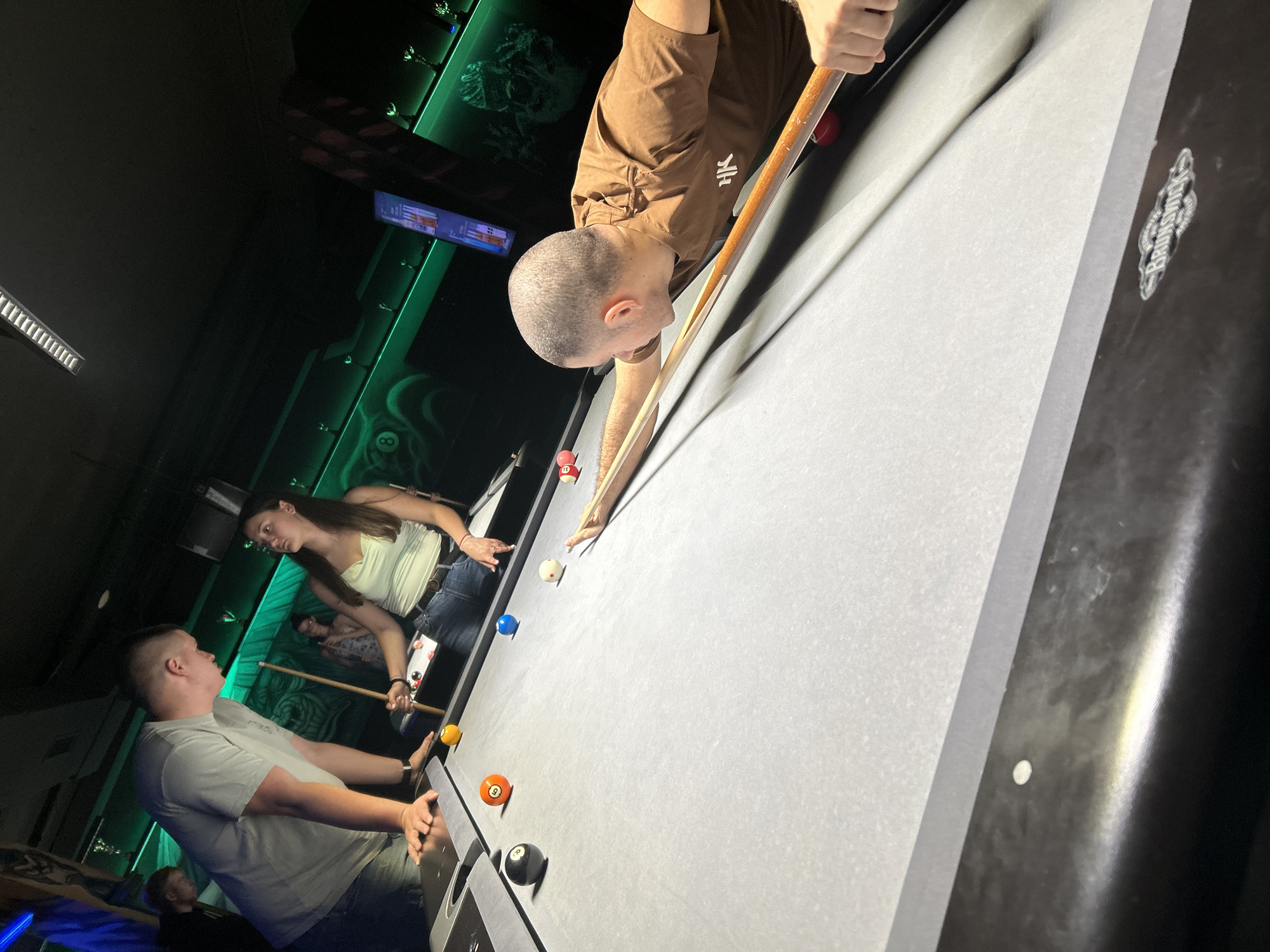 The image captures an active game of pool within the 8 Ball Club, located in Sofia, Bulgaria. Four individuals are present around a Brunswick pool table. In the foreground on the right, a man with a shaved head, wearing a brown t-shirt with a white "HK" logo, is positioned over the table, aiming a shot with a pool cue at a white cue ball. Several solid-colored pool balls are scattered across the light grey felt surface of the table, including an 8-ball, a 5-ball, a 1-ball, a 2-ball, and a partially visible red ball.
To the left of the table, a man in a light grey t-shirt and dark jeans leans with his hands on the table, observing the game. Adjacent to him, a woman with dark hair, wearing a light green tank top and dark jeans, holds a pool cue vertically and watches the shot. A fourth person is partially visible in the dim background on the far left.
The environment is characterized by low lighting and distinct green LED accent lights running along the walls and shelves. The walls feature dark surfaces with illuminated green sections showcasing various decorative elements, including skull motifs and abstract swirling patterns. Trophies or similar items are displayed on the green-lit shelves. A digital display screen, possibly showing a menu or game information, is visible on the right wall. A long, rectangular light fixture is present on the ceiling.