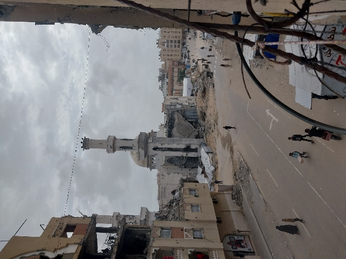 The image depicts a street scene in Khan Yunis, Palestinian Territory, under an overcast sky. In the mid-ground, a severely damaged mosque with a white dome and a tall minaret stands amidst extensive rubble. Its main structure shows significant breaches, exposing interior elements. Flanking the street, multi-story buildings exhibit heavy structural damage, with exposed rebar, missing facades, and debris piles at their bases.
The paved road in the foreground and mid-ground features a solid white lane divider and a painted white turn arrow. The road surface is partially covered with dirt and small debris. Approximately 10-15 individuals are visible on the street and sidewalks. In the lower left, two figures, one wearing a dark abaya, walk along the damaged sidewalk. Closer to the center, three younger individuals walk on the road, one carrying a light-colored object. Further down the street, several other people are dispersed, walking amidst the rubble and surviving structures. One individual is seen operating a motorcycle or scooter in the lower right foreground, partially obscured by bundled wires and metal fragments extending from a damaged structure on the right.