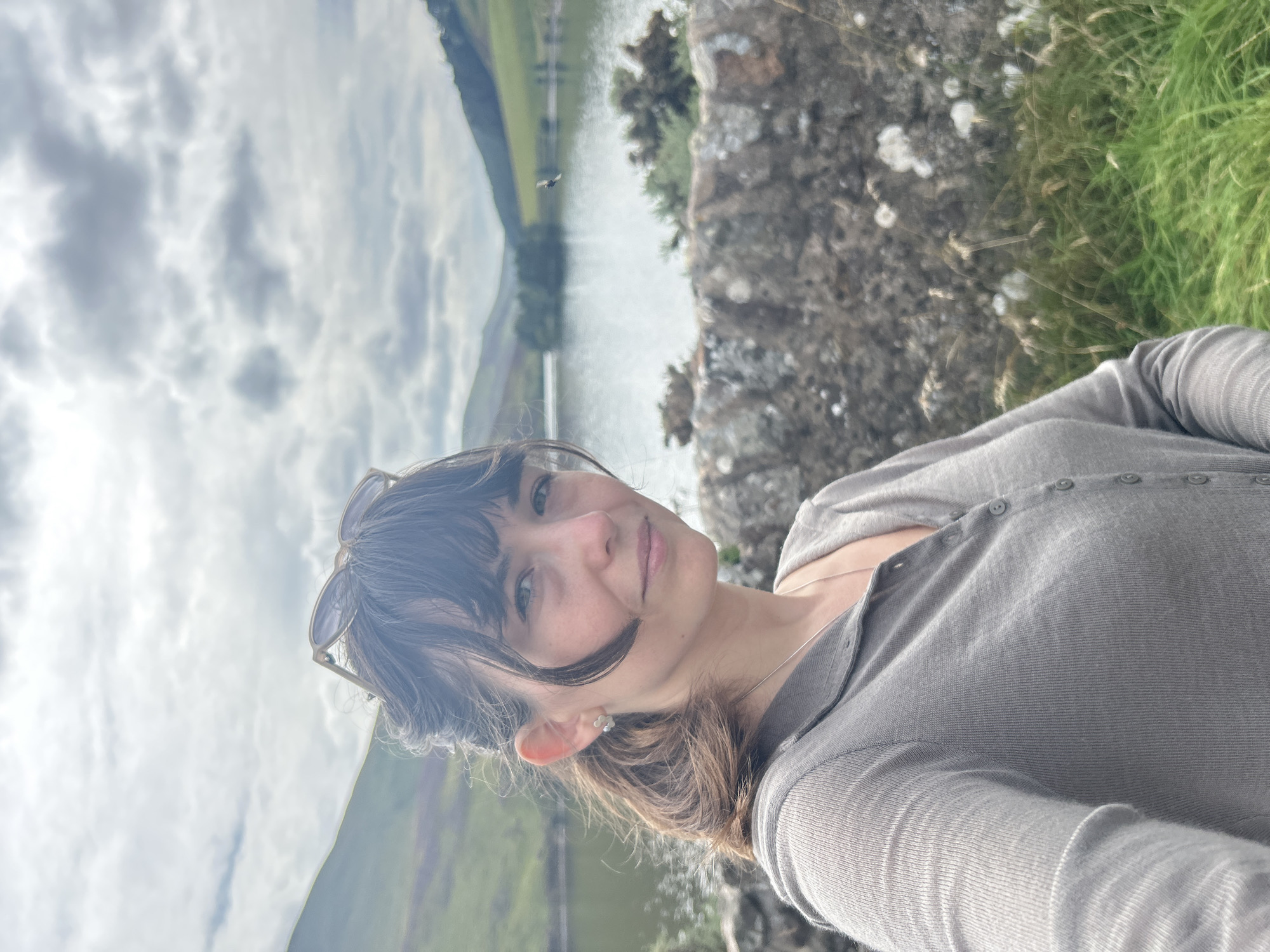 A young woman with dark hair, wearing a grey cardigan and a flower-shaped earring, is standing outdoors in front of a body of water and rolling green hills. She is looking towards the camera with a slight smile. In the background, a stone wall and tall grass are visible. The sky is cloudy. The location appears to be a rural landscape, likely near Penicuik, United Kingdom, given the context.