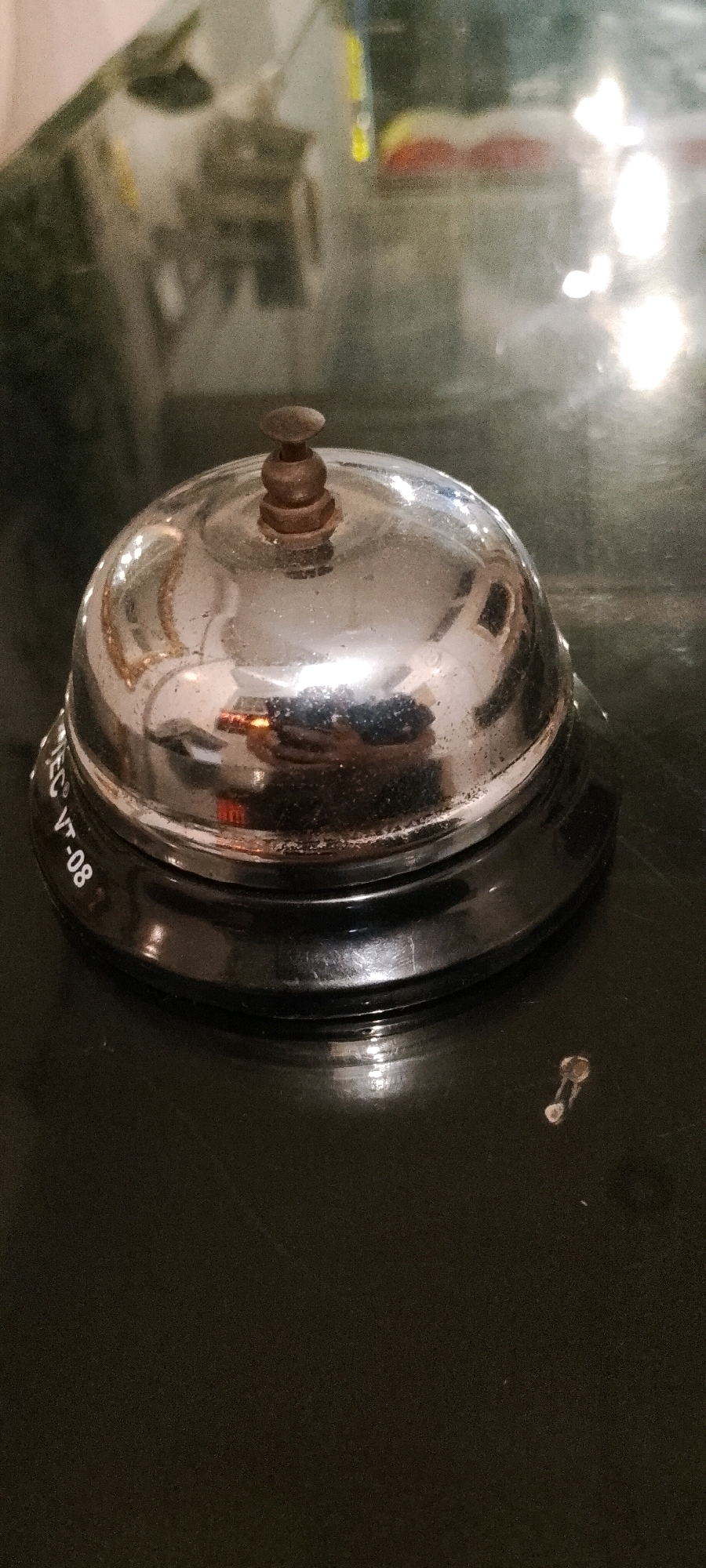A close-up, eye-level, vertical shot shows a chrome-plated, dome-shaped service bell with a small, brown, wooden knob on top, resting on a dark, glossy surface that reflects the surrounding environment, including what appears to be a blurry image of people and lights in the background. Text is visible around the base of the bell, reading "VTEC VT-08". A small, light-colored object, possibly a paperclip or pin, is visible on the surface to the right of the bell. The overall scene suggests a retail or service counter in an indoor environment within the City of Medan, Indonesia.