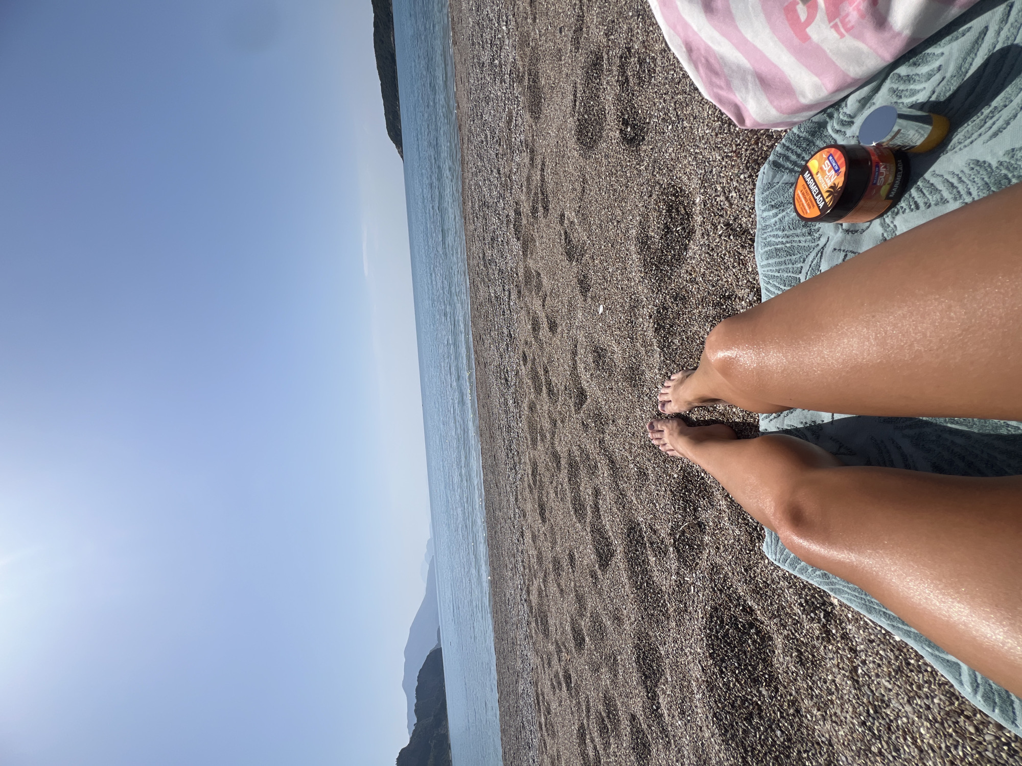 The image provides a first-person perspective of someone relaxing on a pebble beach in Prijevor, Montenegro. The primary subjects are the person's tanned legs and bare feet, stretched out on a light blue/green towel in the foreground.
Adjacent to the legs, an open jar of "OLIVAL SUN PROTECTION MARMELADA" tanning product is visible on the towel, featuring text like "MARMELADA" and "SUN PROTECTION." A pink and white striped bag is partially visible in the upper right corner.
The scene depicts a sunny day at a coastal location. The dark pebble beach stretches towards the calm, blue sea, which occupies the middle ground. In the distance, a range of green mountains rises from the water, silhouetted against a clear, bright blue sky. The strong light suggests it is midday or early afternoon. The overall atmosphere conveys relaxation and enjoyment of a beach vacation.