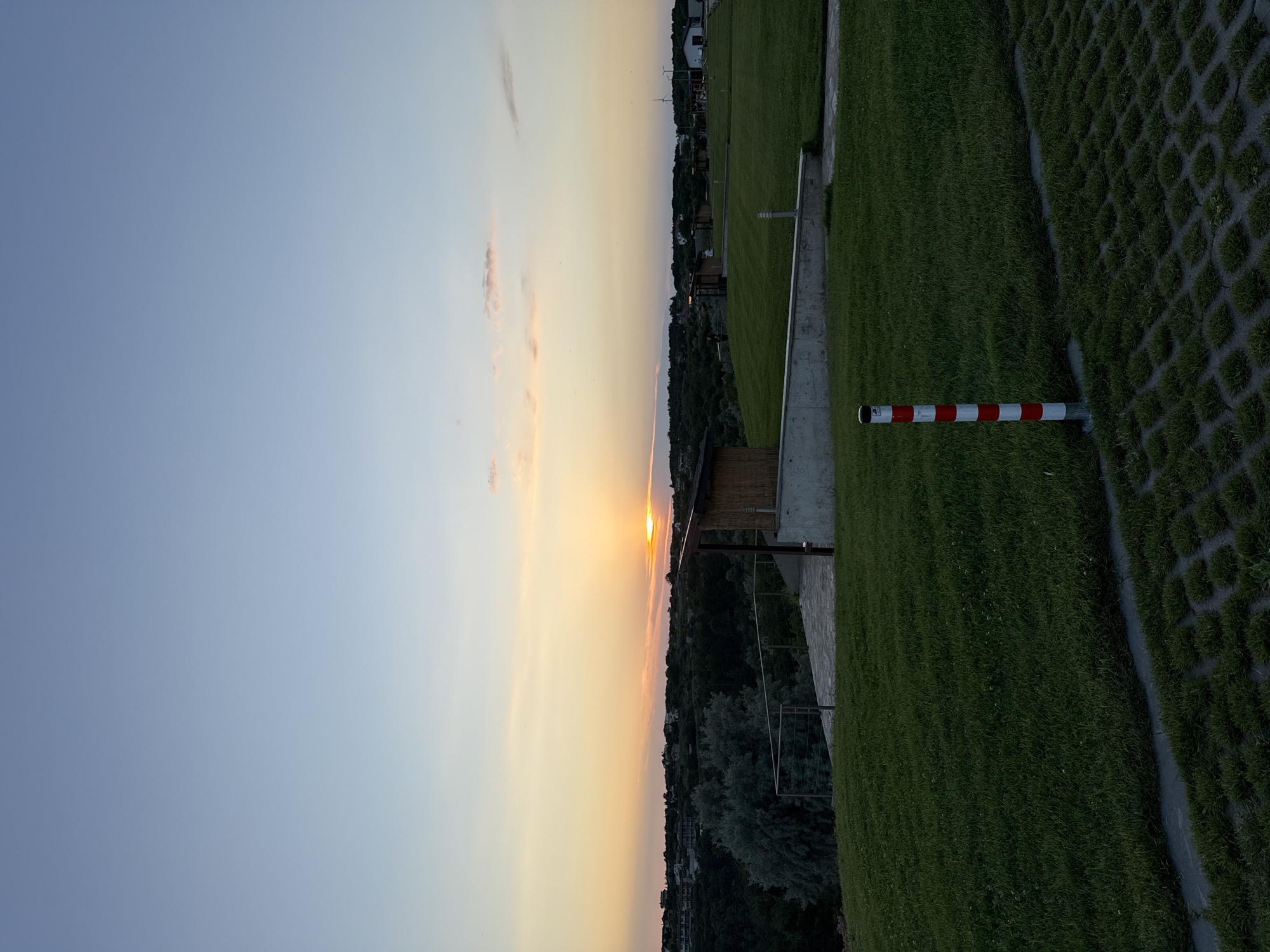 A low-angle, full outdoor shot depicts a scenic view of Topola, Bulgaria at sunset. The sky is a gradient of light blue and orange, with the sun partially obscured by a strip of clouds near the horizon. Below the sky, a lush green landscape stretches out, dotted with trees and buildings. In the foreground, a red and white striped pole stands upright on the grass. A pathway of permeable paving stones is visible in the bottom right corner. The setting appears to be a park or recreational area, with several small, wooden structures scattered across the grassy fields. The overall impression is one of tranquility and natural beauty.