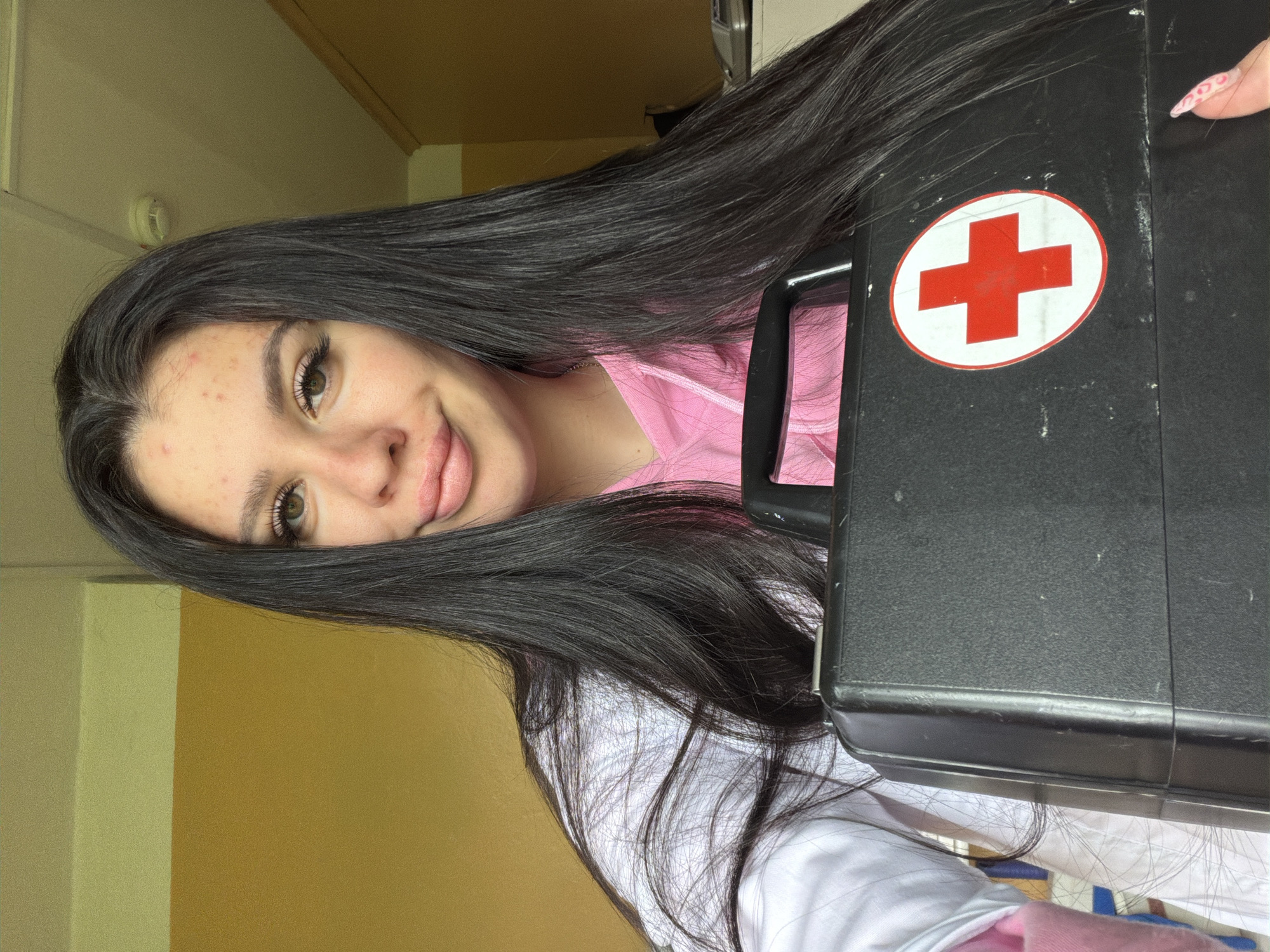 A young woman with long, dark hair and light brown eyes smiles at the camera while holding a black medical bag with a red cross emblem. She is wearing a pink hoodie under a white lab coat. The background appears to be an indoor setting with yellow and beige walls. The image does not provide enough information to determine the specific location within the city of Chumna, Russia.