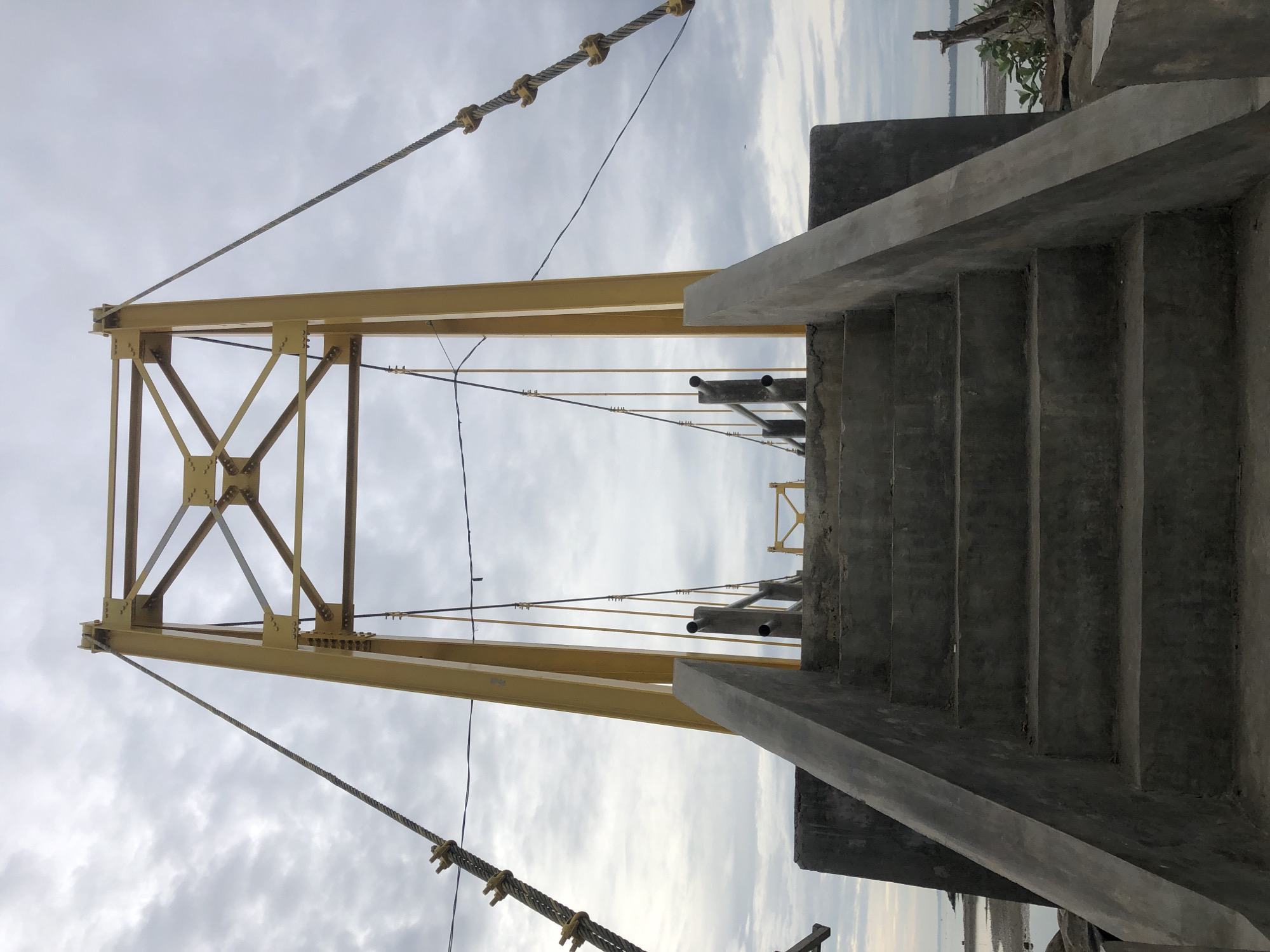 The image shows the entrance to a yellow suspension bridge in Lhokseumawe, Indonesia. Concrete stairs lead up to the bridge's pathway. Thick steel cables, anchored by metal fixtures, extend upwards from the bridge towers. The sky is overcast with clouds. The bridge is situated near a body of water, with a shoreline visible in the background.