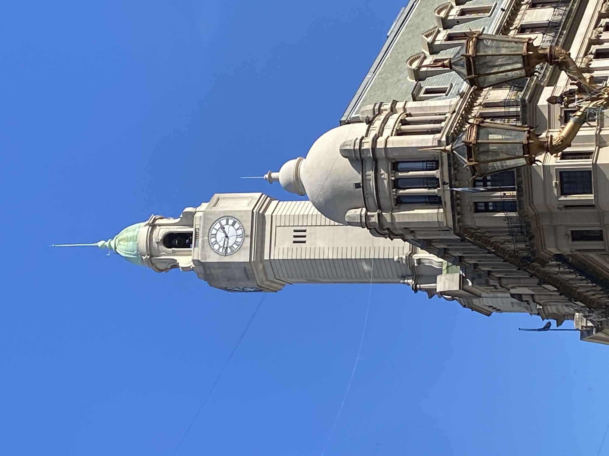 This is a low-angle shot of the Kavanagh Building in Buenos Aires, Argentina. The image focuses on the upper part of the building. The building features a clock tower with a clock face visible. Above the clock tower are architectural details including a dome-like structure and spires. The sky is clear and blue. The photo highlights the building's architecture.