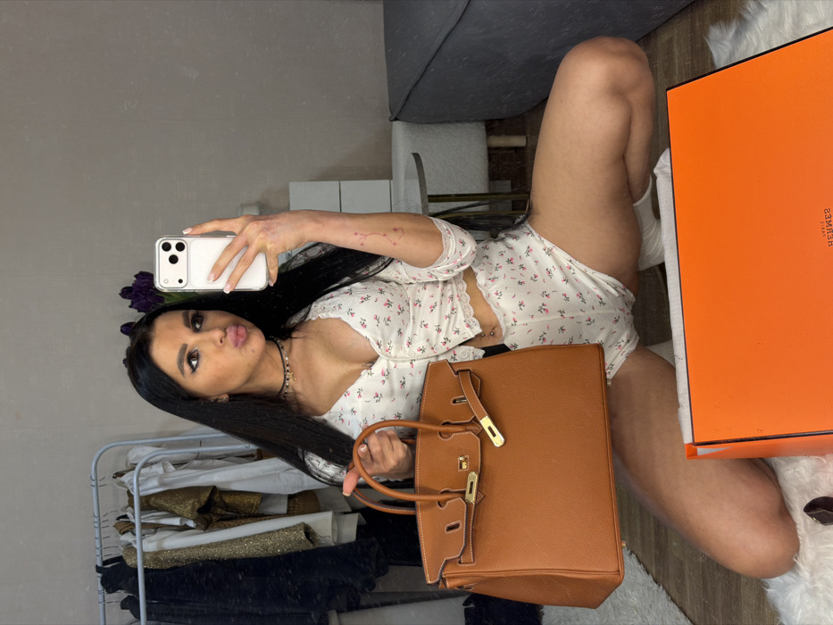 An indoor mirror selfie taken in the city of Svkh Voronezhskiy, Russia, depicts a female subject kneeling on a white shag rug. The subject has long dark hair, prominent eye makeup, and her lips are puckered. She wears a white two-piece set consisting of a short-sleeved, buttoned crop top and matching shorts, both featuring a small floral pattern and white lace trim. A black choker necklace and a navel piercing are visible. Her left arm shows visible skin irregularities. In her right hand, she holds a white smartphone with a clear case, pointed towards the mirror. Her left hand holds a tan/brown leather handbag with gold-toned hardware. An open orange box, displaying the "HERMÈS" logo in mirror writing, rests on the rug in the foreground. The subject wears white socks. The background features a clothing rack with various garments on the left, and a grey upholstered sofa next to a small side table with a dark object on the right. The room's walls are light-colored.