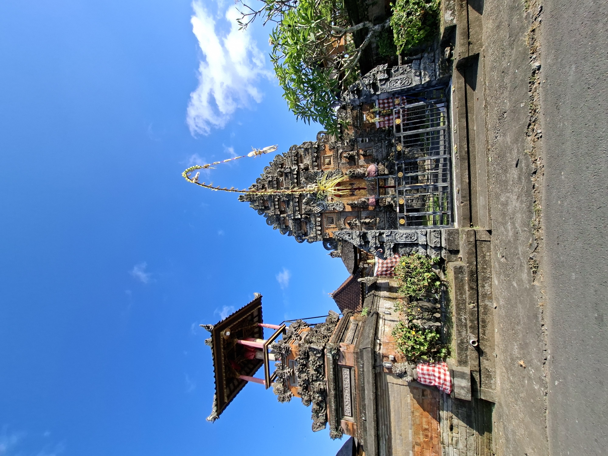 A low-angle, wide-shot captures the exterior of a Balinese temple under a clear blue sky with scattered white clouds. The temple features intricate stone carvings, multiple tiered roofs, and is adorned with red and white checkered fabric. To the left, a tall, ornate tower with a canopy roof stands prominently. In front of the temple is a metal gate and a paved area, with a road visible in the foreground. Lush green trees are present to the right of the temple. The scene is located in Blahbatuh, Indonesia.