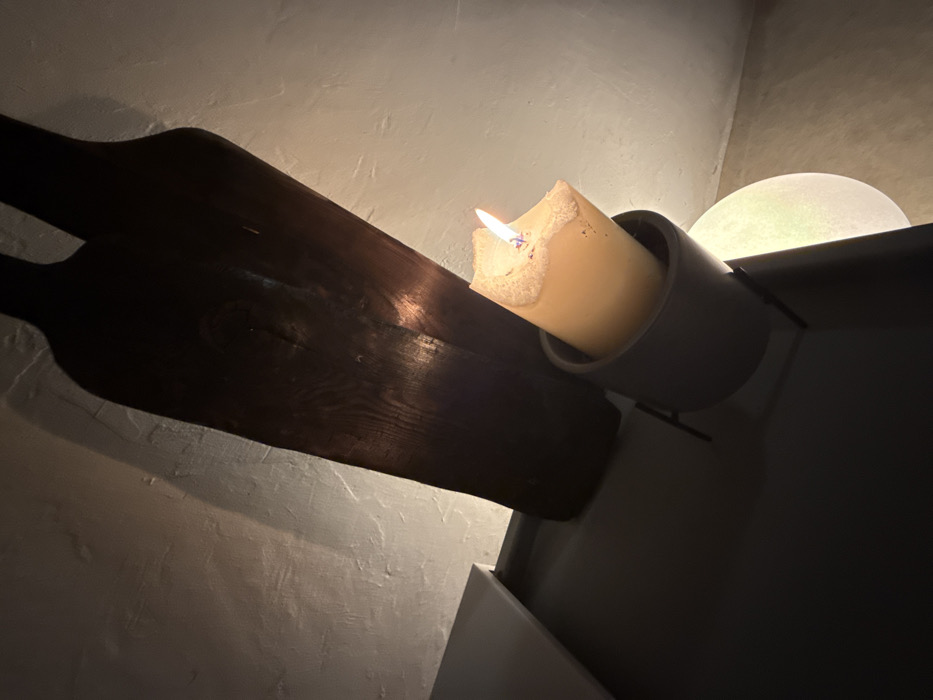 The image depicts an indoor scene in Pryaporets, Bulgaria. A thick, off-white columnar candle with a burning wick and a bright flame is centrally positioned within a dark gray, cylindrical holder. The candle's upper edge exhibits melted and slightly bubbled wax. This holder is mounted on a slender, dark metallic support structure, which extends onto a flat, dark, matte surface in the lower foreground. In the background, two dark, vertically oriented, wood-grained objects resembling stylized bottles or paddles are affixed to a textured, light-colored wall. The candle flame provides the sole direct illumination, casting a warm light and creating prominent shadows from the wooden objects on the wall. A subtle, circular diffused light source is partially visible at the lower right, emanating a soft glow.