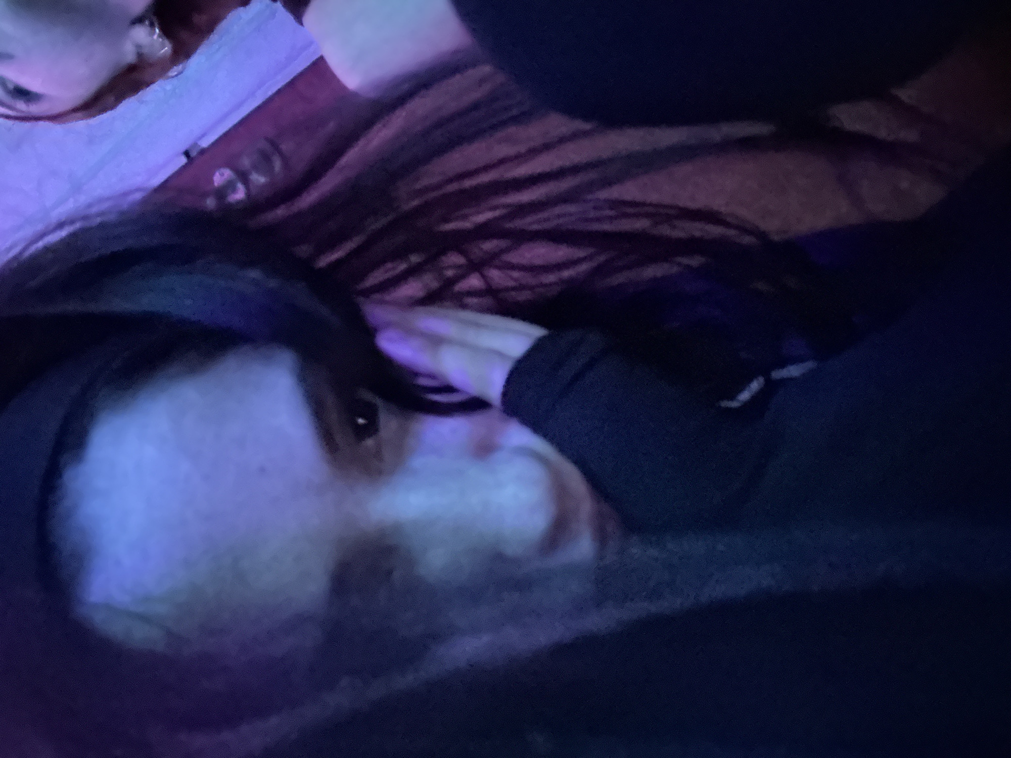 A selfie appears to have been taken indoors with the subject lying down. The subject has dark hair and pale skin. Another person is partially visible in the upper left corner. The location is unknown.