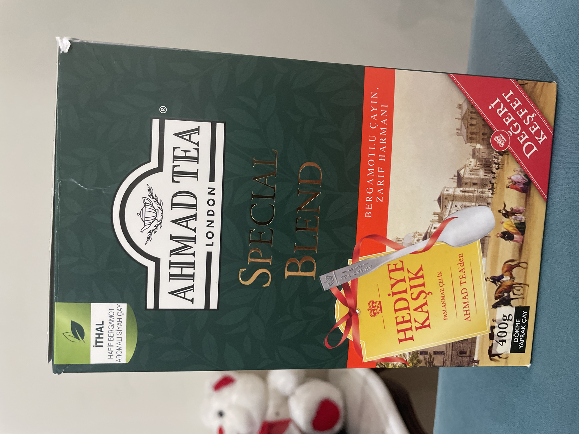 The image shows the front of a dark green tea box for "Ahmad Tea Special Blend" with a light green label in the upper left corner indicating "İTHAL" (Imported), "HAFİF BERGAMOT AROMALI SİYAH ÇAY" (Light Bergamot Flavored Black Tea). The box features a gold foil logo and text. A yellow tag with red ribbon reads "HEDİYE KAŞIK" (Gift Spoon) and "PASLANMAZ ÇELİK" (Stainless Steel). Below this, text says "AHMAD TEA'den" (From Ahmad Tea). There is a depiction of a town square with people and horses. In the lower left corner, "400g DÖKME YAPRAK ÇAY" (400g Loose Leaf Tea) is visible. A red ribbon in the lower right corner says "DEĞERİ KEŞFET" (Discover the Value). The box is placed on a blue, textured surface. The overall context does not provide any specific information about the city of Kadıköy, Turkey, or any elements that can be used to pinpoint the location within Kadıköy.