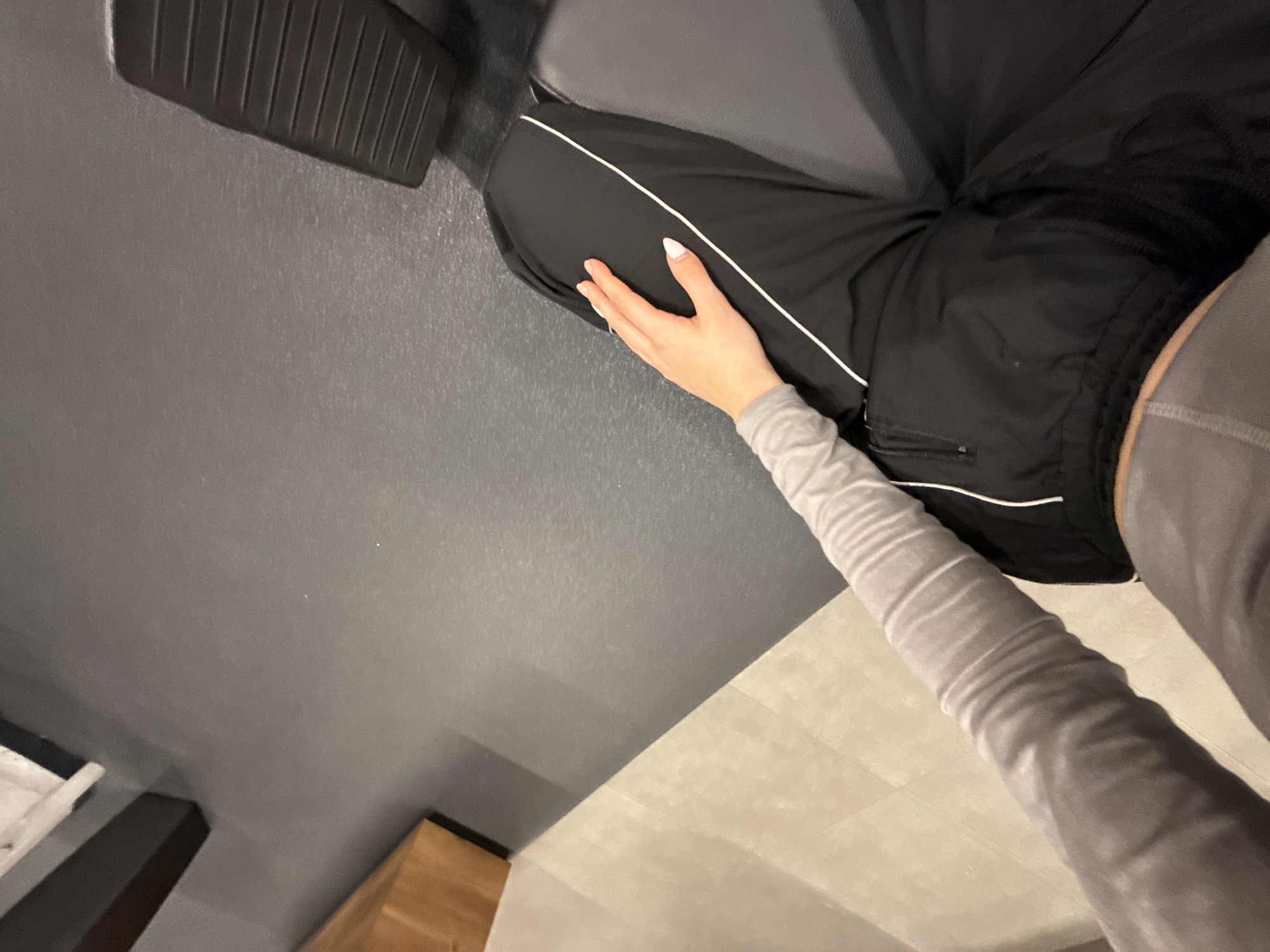 A medium shot depicts a person seated on a black surface with a textured footrest, possibly a gym bench or exercise equipment. The person wears black athletic pants with a white stripe detail and a long-sleeved gray top. The left hand rests on the person's thigh. The background consists of a dark gray wall and a light-colored floor, with glimpses of a wooden cabinet and white molding near the bottom. The image is rotated counterclockwise. It is not possible to determine the location within Sofia, Bulgaria from the given information.