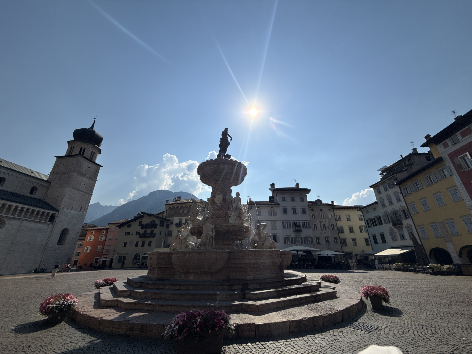 This media file captures a sunny day in a European city square, likely Trento, Italy. The scene features a large, ornate fountain as the central subject, with a statue of a figure at its apex. Surrounding the fountain, cobblestone paving leads to colorful, multi-story buildings with a distinct architectural style, suggesting an older European city. A tall church tower with a dark, domed roof is visible to the left, adding to the historical atmosphere.
The time of day is indicated by the bright sun high in the clear blue sky, casting strong shadows and creating lens flares, typical of midday. The weather appears to be warm and clear, with only a few wispy clouds present. The square is relatively populated with a few people strolling or sitting, creating a relaxed, everyday atmosphere. Potted flowers with pink and white blooms are strategically placed around the fountain, adding a touch of color and life to the urban landscape.
The overall mood is peaceful and inviting, showcasing the charm of a European piazza. The image focuses on the interplay of light and shadow, highlighting the textures of the stone fountain and buildings. No visible text is present in the image.