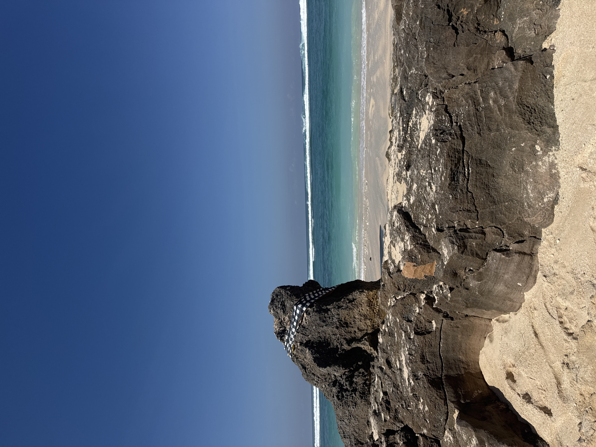 This image depicts a serene coastal landscape with a clear blue sky, turquoise ocean, sandy beach, and prominent rock formations. The upper two-fifths of the frame are dominated by a vast, clear blue sky, slightly lighter towards the horizon. Below the sky, the ocean exhibits a gradient of turquoise and aqua hues, with several parallel lines of white-capped waves breaking in the mid-distance and closer to the shore. A narrow strip of light beige sand forms the immediate shoreline where the waves dissipate.
In the foreground, a large, dark brown and black rock formation with a rough, weathered texture occupies the lower portion of the image, extending from the bottom edge towards the center. Sand is visible at its base and within some crevices. To the left of the frame, a distinct, large, dark grey rock stands upright, its irregular shape silhouetted against the ocean. A black and white checkered cloth, resembling a sarong or ceremonial fabric, is draped over the top and down the right side of this prominent rock. No people, animals, or additional man-made structures are visible in the scene. The lighting suggests bright daylight, casting subtle shadows on the rocks.
Location: Unknown, Unknown.