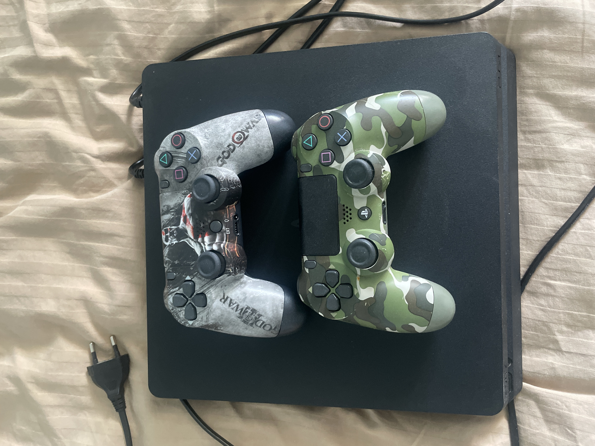 Two Playstation 4 controllers are shown placed on top of a black Playstation 4 console. The top controller has a "God of War" theme with an image of Kratos. The bottom controller has a camouflage pattern in shades of green and brown. The console and controllers are resting on a beige, textured surface, likely a bedspread. A black power cord with a European-style plug is visible to the left of the console, disconnected. Black cables also extend from the console to the right and disappear out of frame. The image appears to be taken indoors in natural light. No people are present in the scene. The location in Kyiv, Ukraine is not discernible from the image.
