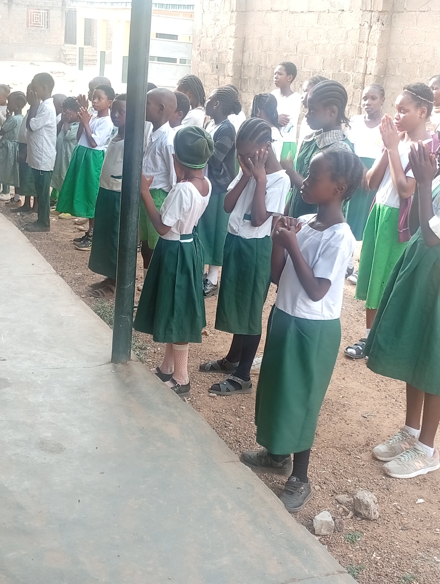 In Narayi, Nigeria, a group of approximately 20 children stand in rows outdoors, likely at a school. They are dressed in school uniforms consisting of white shirts and green skirts or trousers. Most of the children are Black. Many have their hands clasped in front of them, suggesting they may be praying or at attention. Some children have their hands covering their faces. A metal pole stands among them. The ground is a mix of concrete and dirt. Buildings are visible in the background. The overall lighting suggests it is daytime.