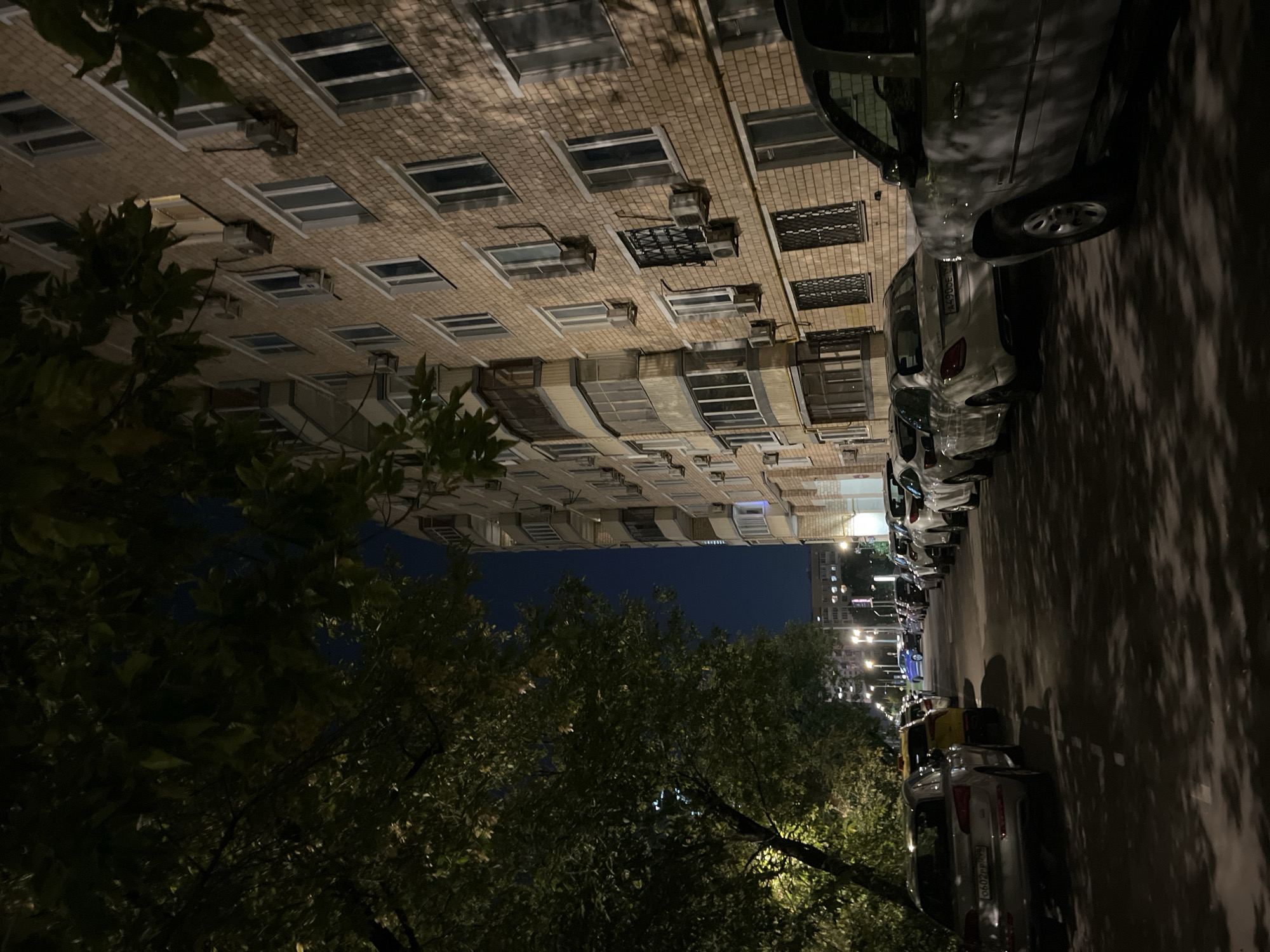 The image depicts a street scene at night in Moscow, Russia, with a residential building on the right and a line of parked cars on the left. The building is constructed of light brown brick and features numerous windows and several air conditioning units mounted on its facade. A section of the building has balconies with glass enclosures. Trees with dense foliage overhang the street from the left, casting shadows on the road and the parked cars.
The parked cars are lined up along the side of the street. The vehicle in the foreground on the right is a dark-colored SUV. To its left, a white sedan is parked, followed by other cars of various colors, including a yellow car and several white and silver sedans and SUVs. The street is paved and appears to be an urban residential area. The sky is dark, indicating nighttime. Streetlights are visible in the background, illuminating parts of the street and other buildings. No people are visible in the frame.