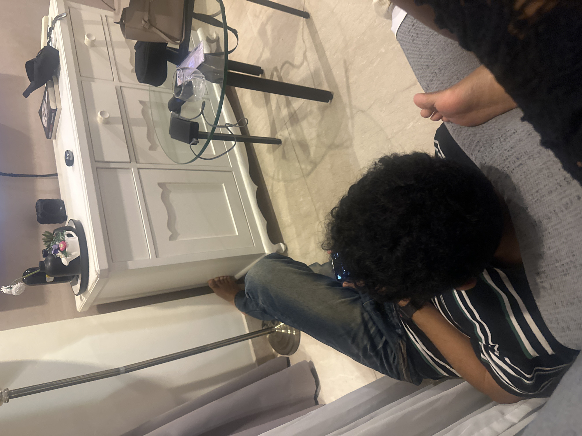 The image shows a person with curly dark hair sitting on a light grey couch, looking down at a mobile phone. The person is wearing a blue, striped shirt and blue jeans. A bare foot is visible near the top of the couch. The floor is a light, neutral tone. Next to the couch is a white cabinet with drawers. On top of the cabinet are various items, including bags, books, and decorative objects. A small, round glass table sits in front of the cabinet. The room appears to be inside an apartment, possibly in a residential area. There is no definitive evidence to suggest this is in Central Jakarta, Indonesia.