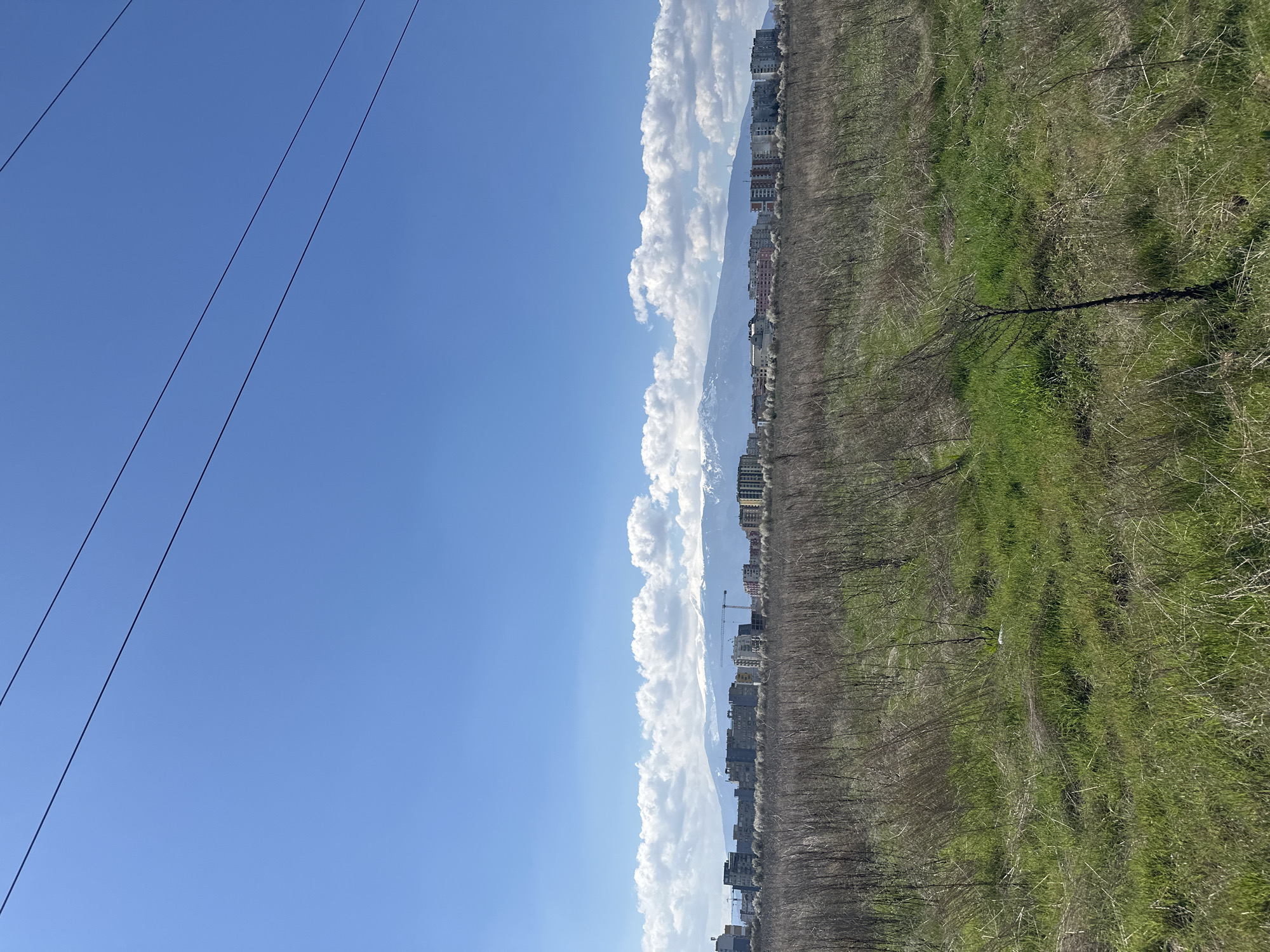 The image depicts an outdoor scene in Sofia, Bulgaria, characterized by a combination of natural and urban elements under a bright, clear sky. In the left foreground, electrical power lines traverse the blue sky. Adjacent to this, a line of cumulus clouds extends along the horizon, partially obscuring a mountain range in the background. In the center and right foreground, a field transitions from dry, barren vegetation to patches of green grass. In the background, buildings form the urban landscape of Sofia. A construction crane is visible among the buildings.