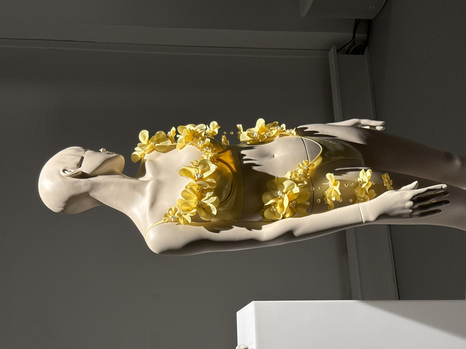 A pale-skinned, bald, female mannequin is displayed in profile, facing right, within a dimly lit interior space in Paris, France. The mannequin is adorned in a bright yellow two-piece garment. The top is a bra-style piece with thin shoulder straps, heavily embellished with numerous yellow fabric flowers, each featuring a central white pearl-like bead. These flowers extend from the bra cups diagonally upwards towards the mannequin's right shoulder and neck. The matching yellow high-cut bottom is similarly decorated with yellow flowers and white pearl beads, forming a diagonal arrangement along the mannequin's left hip and upper thigh. Strong directional lighting from the upper left illuminates the mannequin and garment, casting sharp shadows on its right side and the plain dark gray wall behind it. To the far left, a section of a white rectangular pedestal is visible, partially obscuring a small object on its surface. A light-colored baseboard runs along the lower portion of the background wall. The scene suggests a retail, exhibition, or museum environment.