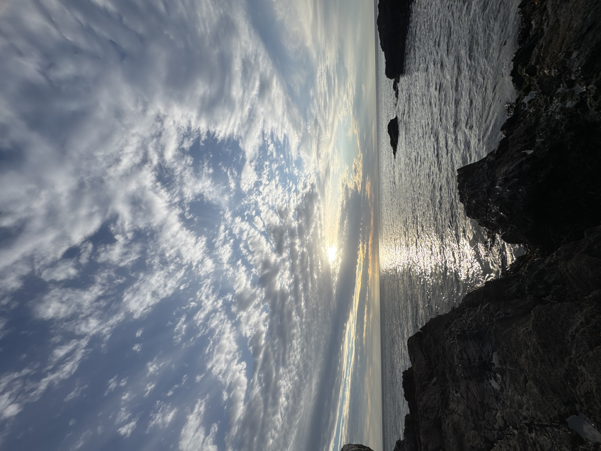 The image shows a coastal scene with the horizon running vertically. The left side of the image depicts a cloudy sky with the sun shining through the clouds. The right side of the image shows the ocean with sunlight reflecting off the surface, and dark rocks in the foreground. The location is Unknown, Unknown.