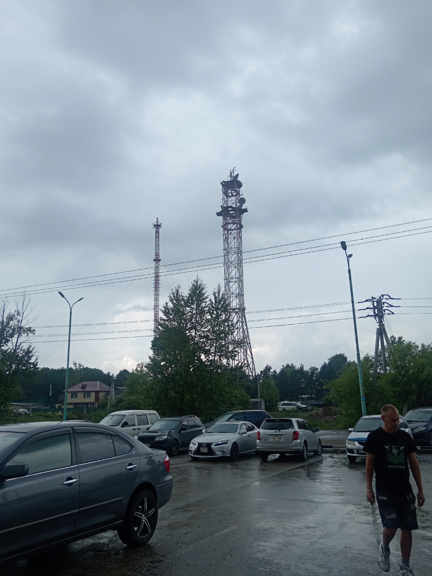 A man in a black t-shirt and denim shorts walks across a wet, asphalt parking lot. In the background, several cars are parked, including a grey sedan, a silver SUV, a white minivan, and a white Lexus. Two tall telecommunication towers dominate the skyline, with one appearing to be taller than the other. Power lines are strung between poles. Trees and a house with a red roof are visible behind the parking lot. The sky is overcast and grey. The scene appears to be in Belogorsk, Russia, possibly during or after rainfall, given the wet pavement.