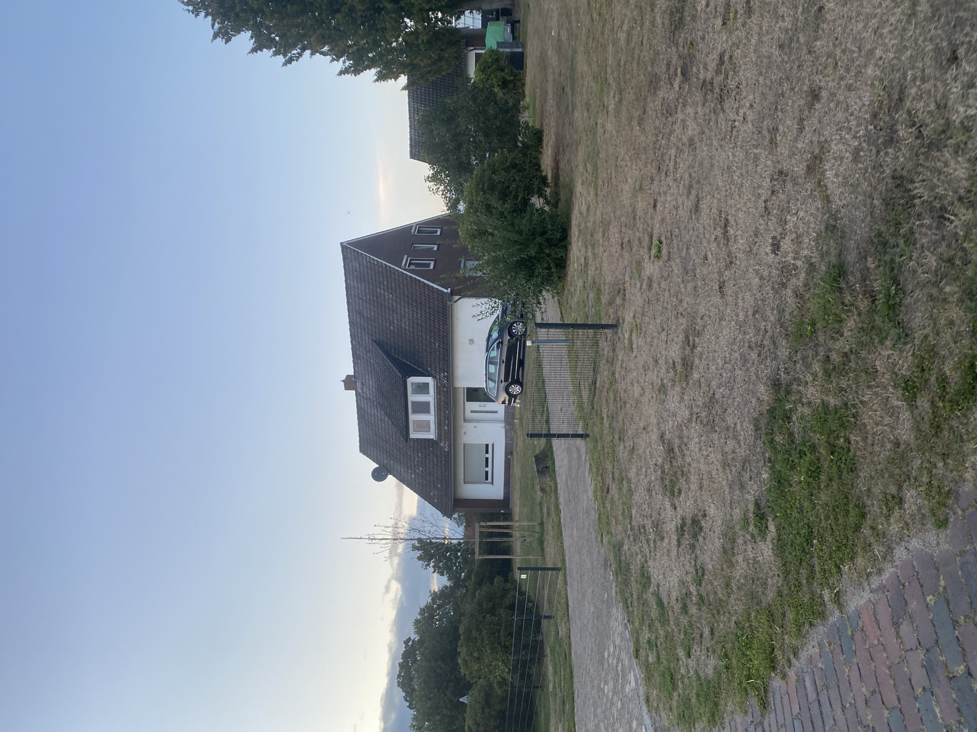 The image depicts a residential scene on a clear day, likely in the evening due to the soft lighting. A two-story house with a dark tiled roof and white siding is prominently featured. The house has multiple windows, including dormer windows on the upper level. A dark-colored sedan is parked in the driveway next to the house.
The property is surrounded by a metal fence. In the foreground, a brick-paved walkway is visible to the left, leading towards the property. The lawn area appears dry and patchy, with areas of brown grass and some scattered green patches. A young tree, supported by wooden stakes, stands in the yard.
To the right of the main house, another building with a similar dark tiled roof is partially visible. This building also appears to be a residence or a garage. Further to the right, tall evergreen trees are present. A green tarp-covered object, possibly a trailer or equipment, is situated near the secondary building.
The sky is a pale blue with some wispy clouds. The overall impression is a quiet, suburban setting in Schwarme, Germany, characterized by typical residential architecture and landscaping. There are no people visible in the image.
