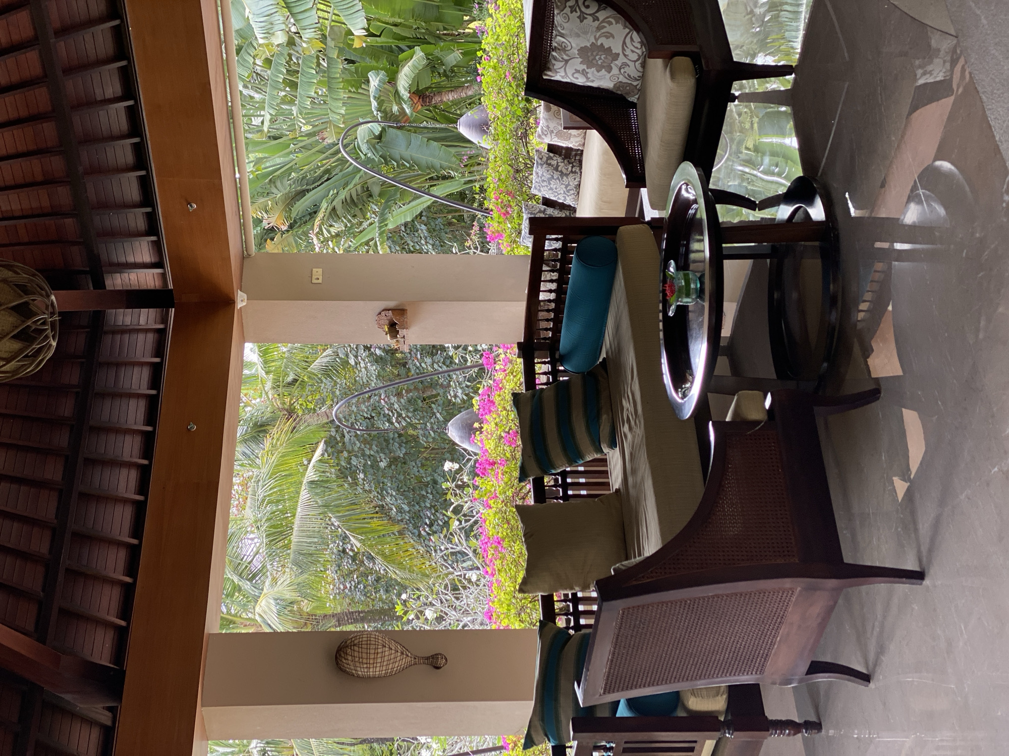 The image shows a covered outdoor seating area with tropical foliage in the background. The scene is furnished with dark wooden chairs and a sofa, all adorned with cushions. A circular coffee table with a glass top sits in the center, holding a small vase with a red flower and green leaves. The floor is tiled with a light gray marble.
To the left, a wooden ceiling structure is visible, and a woven lamp hangs from it. A decorative item resembling a tennis racket made of woven material is mounted on a cream-colored pillar. The background is filled with lush green palm trees and vibrant pink flowers, suggesting a warm, tropical climate. The lighting indicates it is daytime, with natural light illuminating the space. The overall ambiance is serene and inviting, suitable for relaxation.