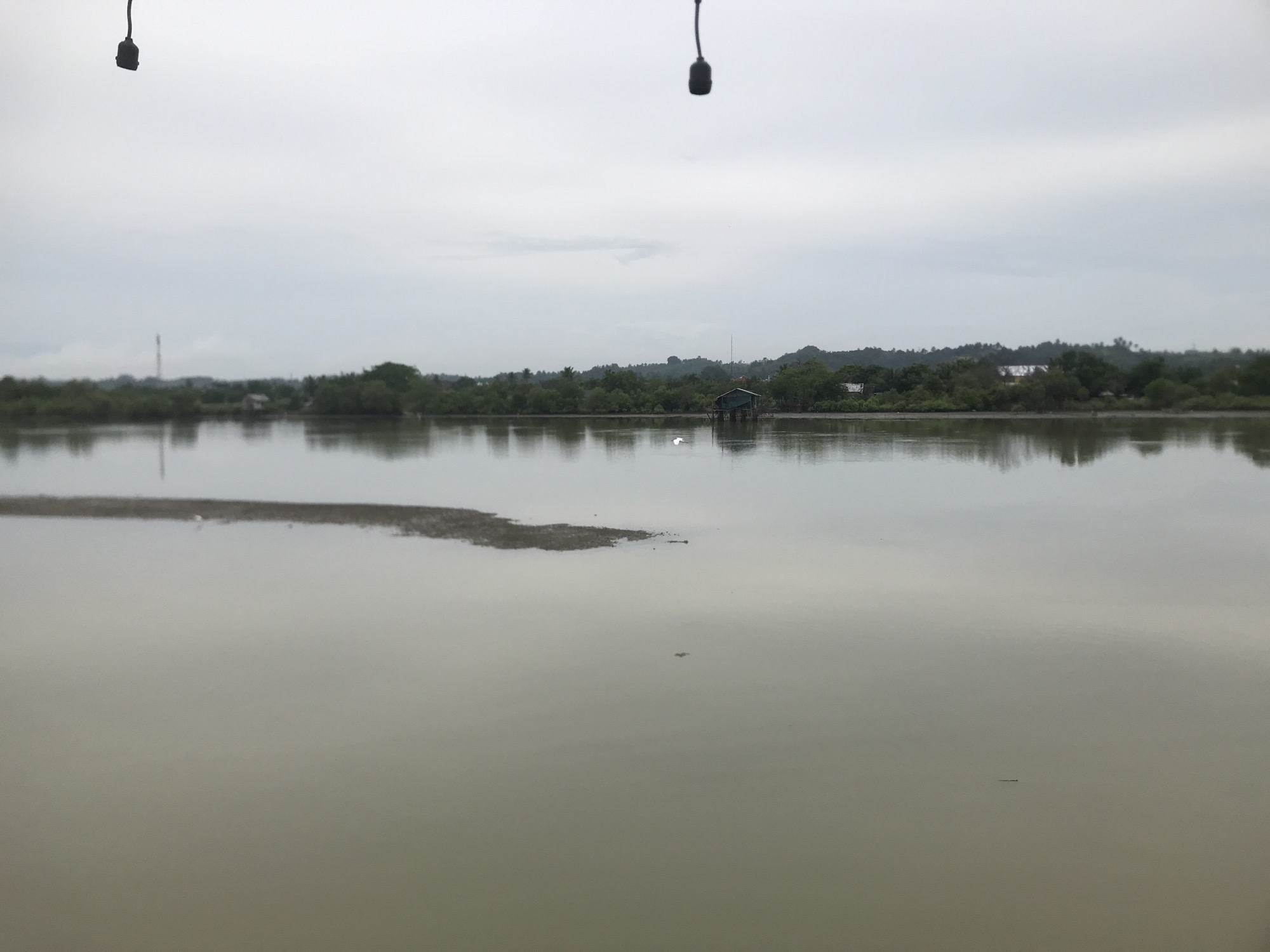 The image depicts a gray and overcast day in Lhokseumawe, Indonesia. A large body of water dominates the scene, its surface relatively still with reflections of the sky and surrounding landscape. In the middle distance, vegetation lines the far bank, interrupted by a small structure on stilts. A small island or sandbar appears in the water closer to the viewer. Two wires with dangling objects are visible in the top corners of the frame, suggesting overhead lines or lighting. No people are visible in the frame.