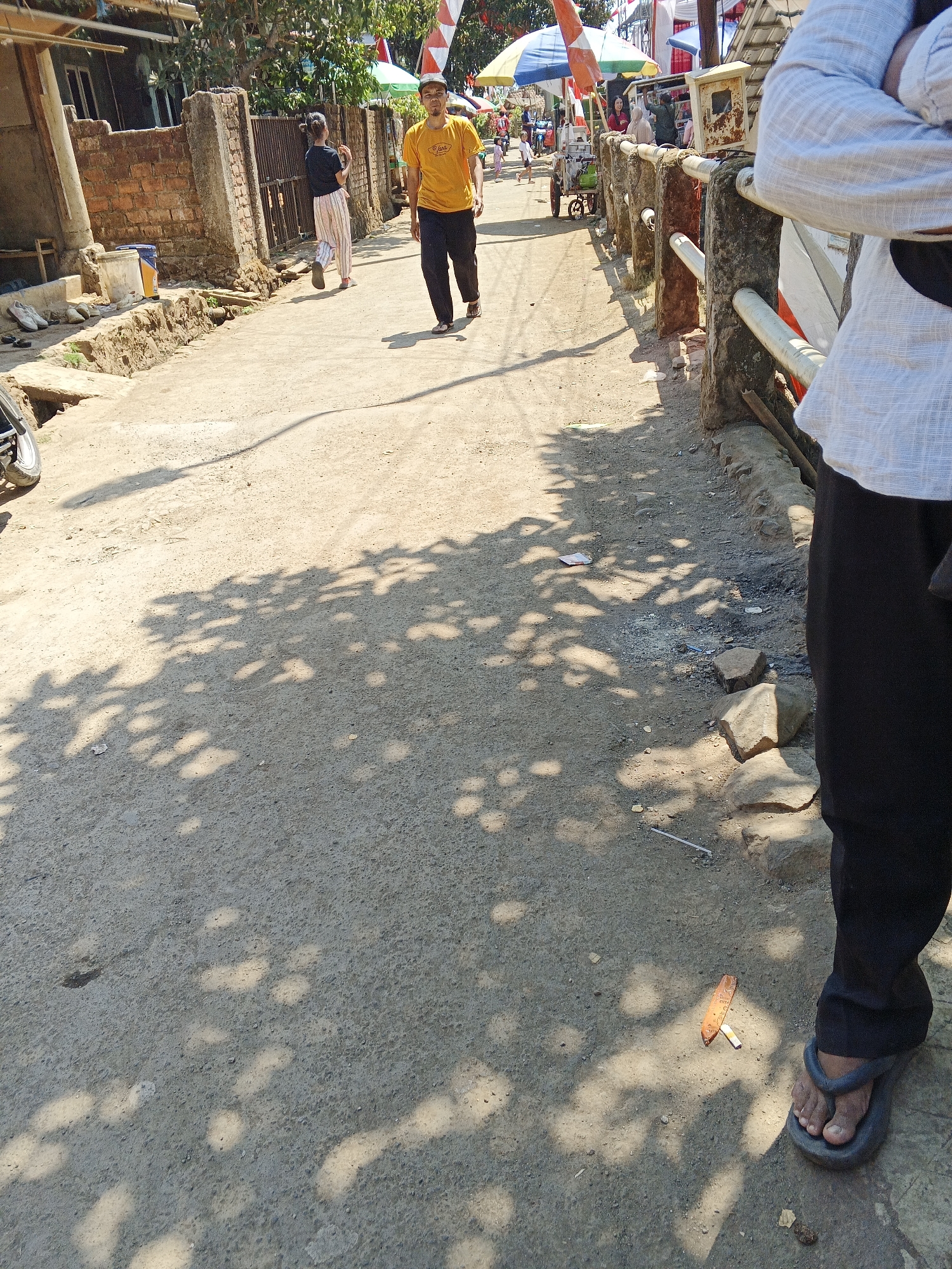 A man wearing a yellow t-shirt and dark pants walks on a dirt road in Sukaharja, Indonesia. To his left, a woman in a black shirt and striped pants walks away from the camera. The road is lined with brick walls and a concrete railing. Trees cast dappled shadows on the ground. In the background, there are buildings and a market with colorful umbrellas. A person wearing a white shirt and black pants is standing on the right side of the image, facing away from the camera. There is a cigarette butt and some trash on the ground near the person in the white shirt.