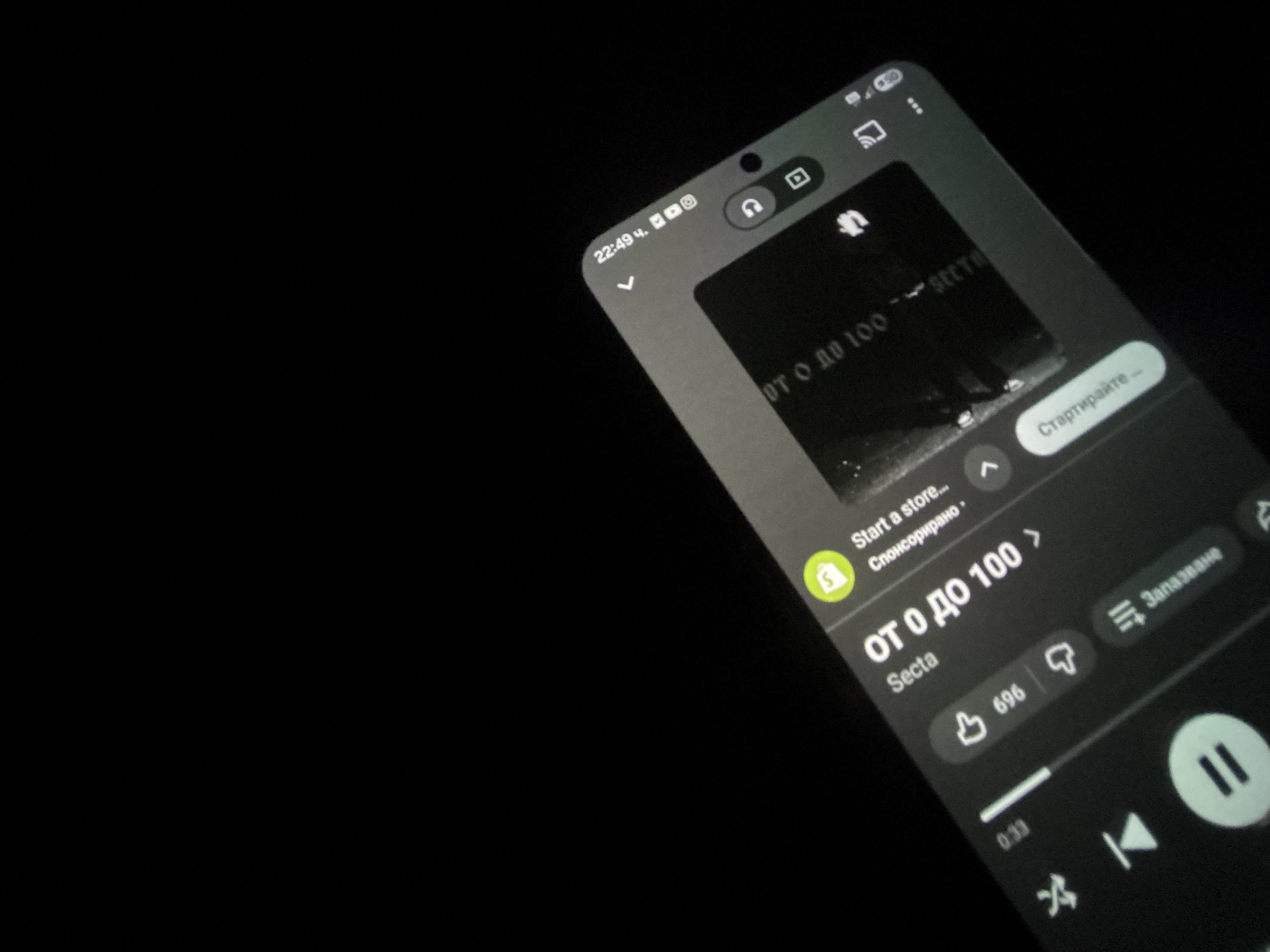 The image shows a mobile phone screen displaying the YouTube Music app. The app is playing the song "ОТ 0 ДО 100" (From 0 to 100) by Secta. The current time displayed on the phone is 22:49. There is a Shopify advertisement present at the top of the screen, titled "Start a store..." The screen shows the thumbs up and thumbs down icon, and other music player controls. The rest of the image is black. There is no indication of Koprivlen, Bulgaria or any other location.