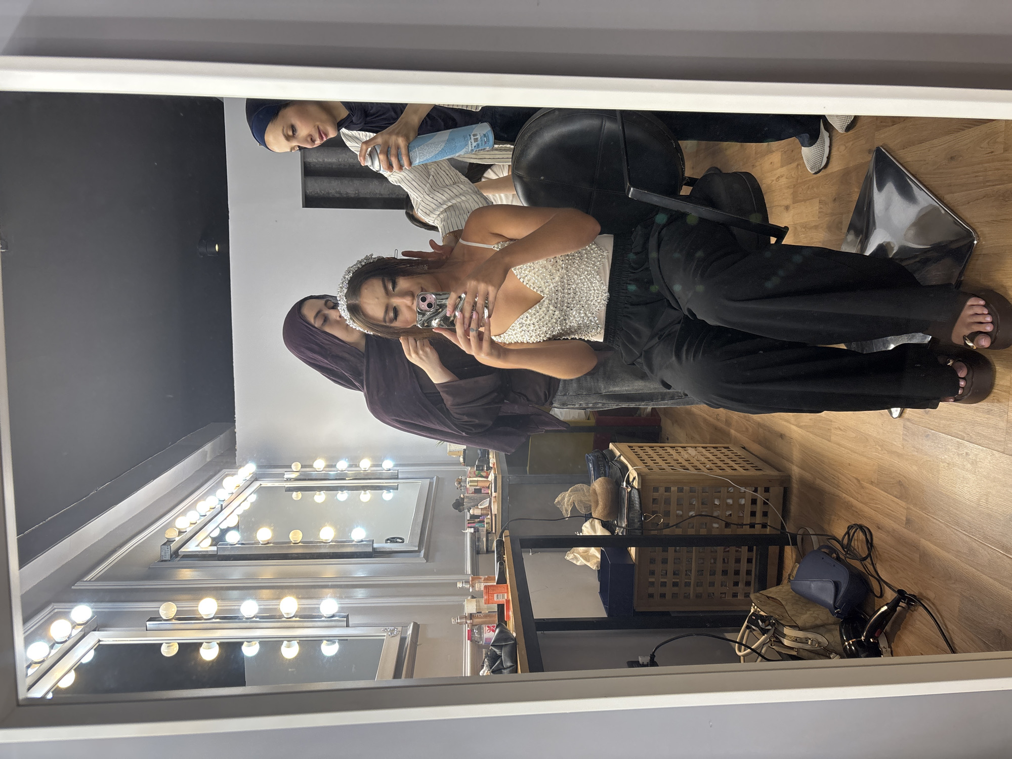 The image captures a reflection in a mirror of a beauty salon environment, likely located in Sweileh, Jordan. In the foreground of the reflection, a young woman sits in a black salon chair, holding a smartphone and taking a selfie. She wears a sparkly white top, black pants, and platform sandals. A tiara is positioned on her head, suggesting preparation for a special event. Two other individuals, presumably makeup artists or stylists, are attending to her. One, wearing a purple headscarf, appears to be adjusting her hair. The other, in a dark headscarf and striped shirt, is holding a can of hairspray. The background of the salon features a vanity mirror with numerous lights, makeup products on a table, and a woven laundry basket containing items. The floor is wooden. The overall scene depicts a moment of preparation and styling.