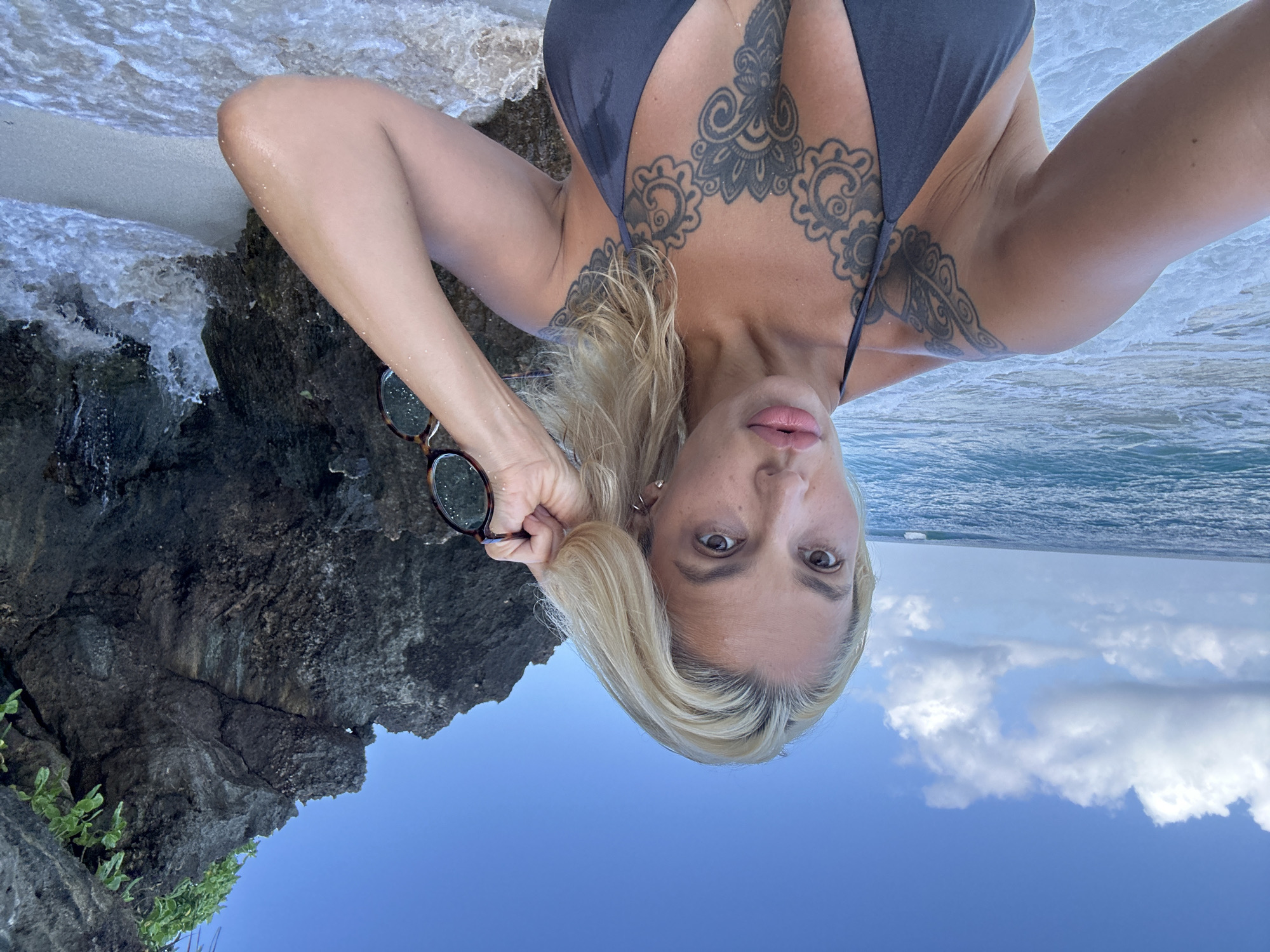 A medium shot photograph captures a blonde woman with light skin, wearing a dark grey bikini top, posing at a beach in Kuta Selatan, Indonesia. She has tattoos on her chest and left shoulder. Her right hand is raised, touching her hair, and she holds a pair of sunglasses with a tortoiseshell pattern in her hand. She looks directly at the camera with full lips and light brown eyes. The background features the ocean with waves crashing on a sandy beach, dark volcanic rocks on the right, and a blue sky with white clouds. The overall impression is that of a selfie taken during a sunny day at the beach.
