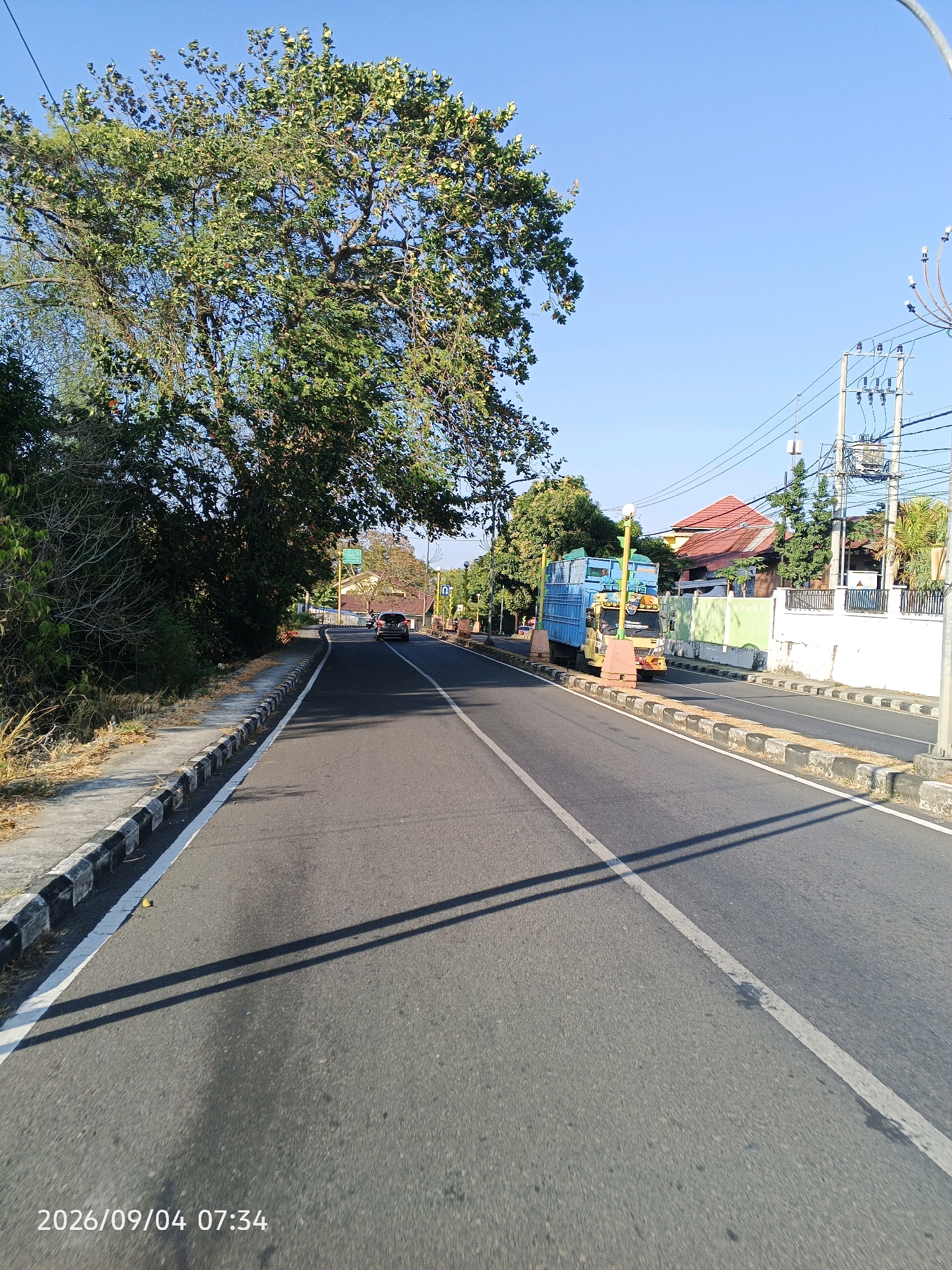 The image captures a sunlit asphalt road in Pare-Pare, Indonesia, as indicated by the timestamp "2026/09/04 07:34". The road is flanked by a raised sidewalk with a black and white striped curb on the left, and a line of trees and vegetation. On the right side of the road, there are residential buildings with red-tiled roofs, a white fence, and several trees. A utility pole with transformers and electrical wires is visible. A blue truck with a yellow cab is parked on the side of the road. A dark-colored car is moving away from the camera in the distance. The sky is clear and blue, and the sun casts long shadows across the road, suggesting it is early morning.