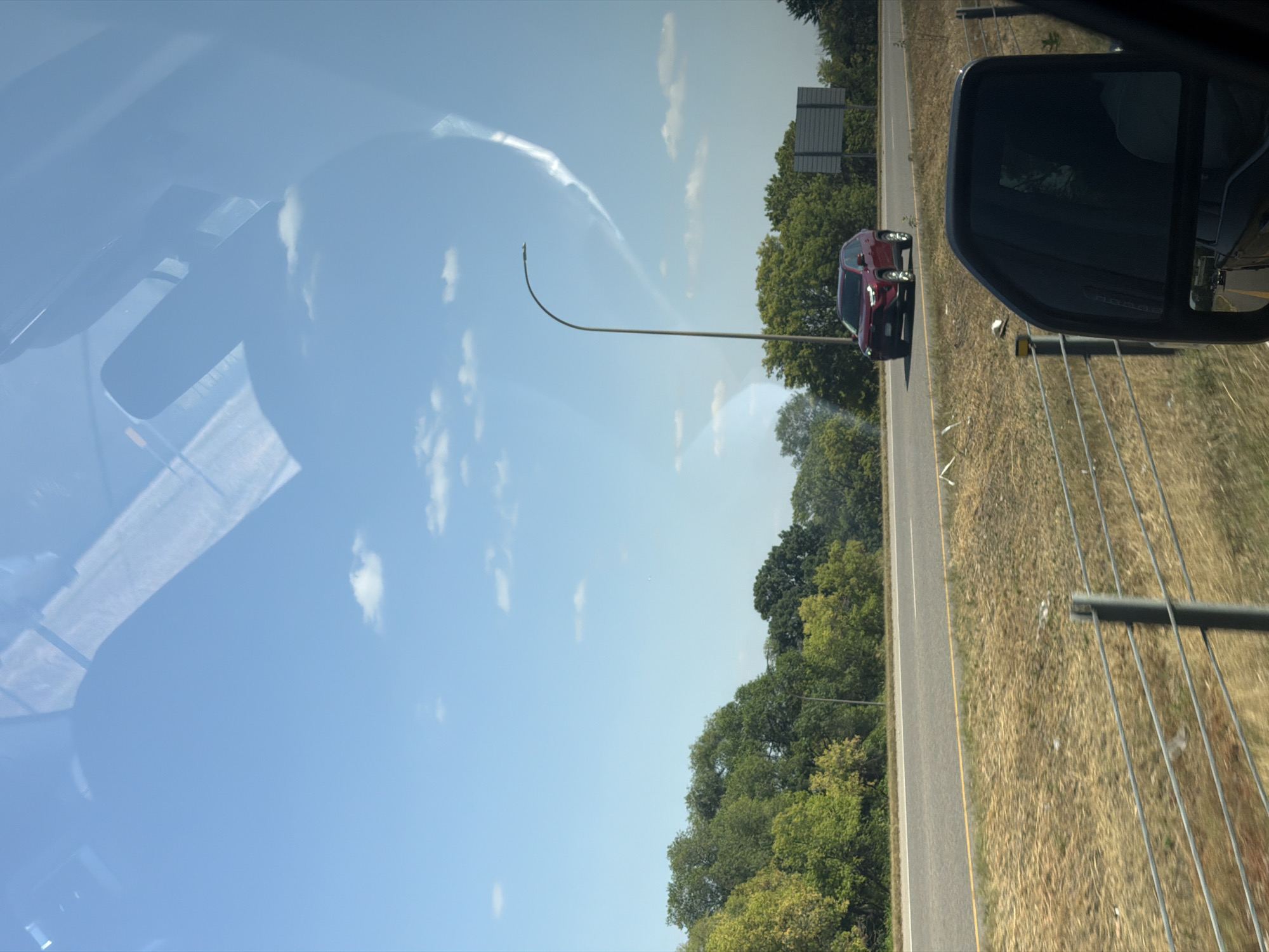 A red SUV drives on a highway in West Saint Paul, Minnesota, United States of America. The SUV is on the right lane of a two-lane road, moving in the same direction as the viewer. Trees line the side of the road on the left, and dry grass is visible on the right. The sky is blue with scattered white clouds and a visible contrail. A street light stands tall on the right side of the road. The image is taken from inside a vehicle, with the side mirror visible in the foreground.