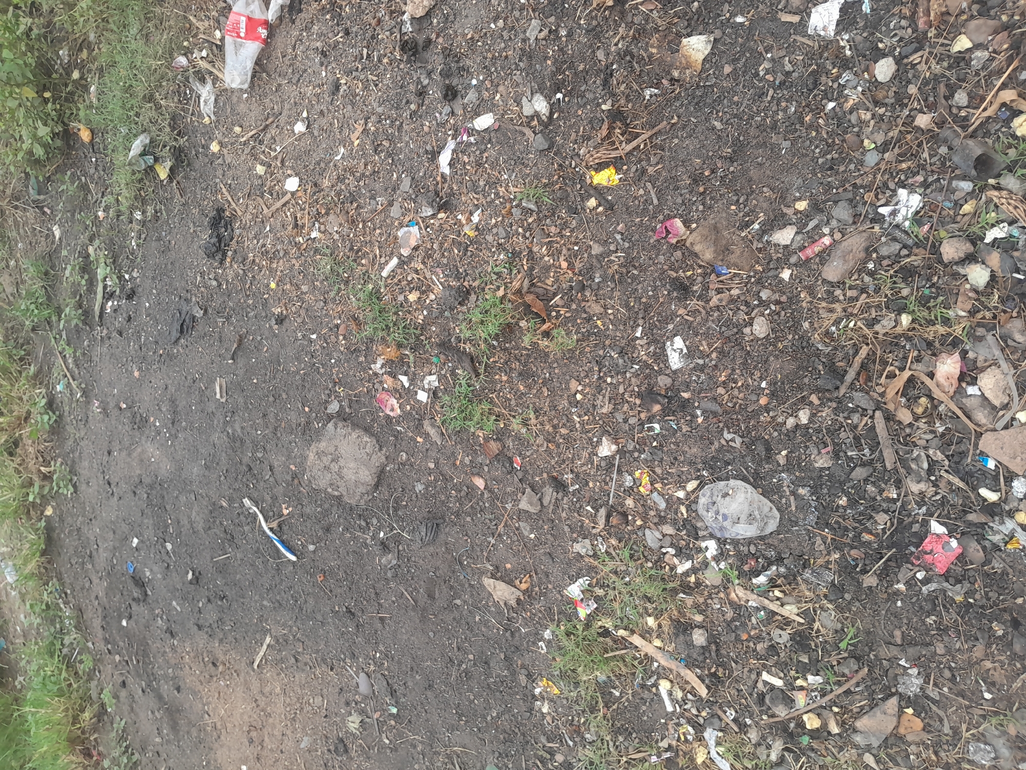 The scene depicts a heavily littered, unpaved ground surface located in Ifo, Nigeria. The ground consists of dark, granular soil interspersed with small stones, pebbles, and sparse patches of green grass. Numerous discarded items are scattered across the area. Visible objects include a partially crushed clear plastic bottle with a red label fragment on the right side, a white and blue plastic toothbrush, multiple fragments of plastic bags and wrappers, torn pieces of colorful paper, and various unidentifiable small debris. Additionally, dried leaves, twigs, and small plant matter are mixed with the soil and litter. A dark, irregular rock or compacted dirt clump is present near the center-left. No individuals or specific actions are depicted.