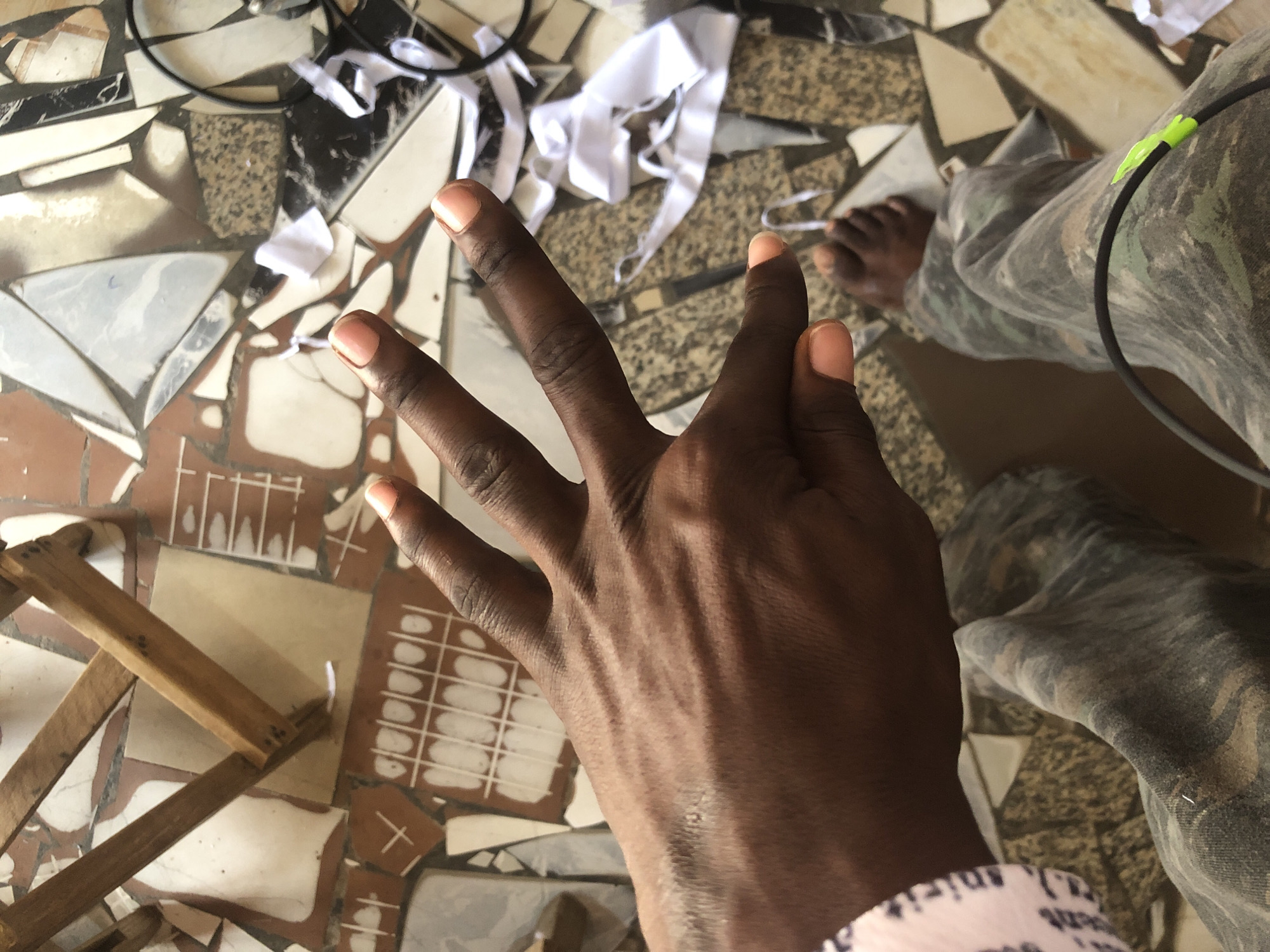 In Ado, Nigeria, a person's right hand with light pink-colored fingernails is held up, palm facing upwards, against a mosaic-tiled floor. The hand is positioned over the person's legs, which are clad in camouflage pants. The floor's tiles are broken and irregularly shaped, with patterns of white, grey, brown, and black. Some white fabric scraps and a black cable with a green tip are also visible on the floor. A wooden structure, possibly part of a chair, is present in the upper left corner of the image.