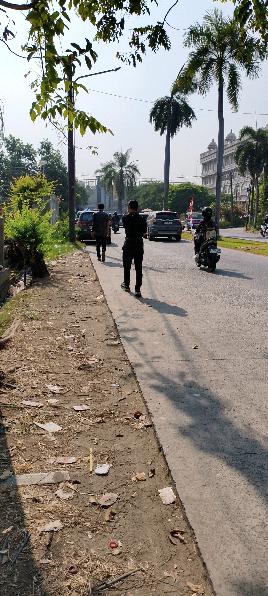 In West Jakarta, Indonesia, on a sunny day, a group of individuals are visible on a street. In the foreground, a person in black attire is standing on the sidewalk, facing away from the camera and appearing to be on a phone call. Further down the sidewalk, another individual in dark clothing walks away from the camera. A motorcycle rider in black with a helmet is on the street, moving away from the viewer. Several cars are also present on the road. The environment consists of a paved road and sidewalk, with a dirt and debris-covered area adjacent to the sidewalk. Palm trees and other foliage are visible, along with the facade of a light-colored building in the background. The sky is clear and blue.
