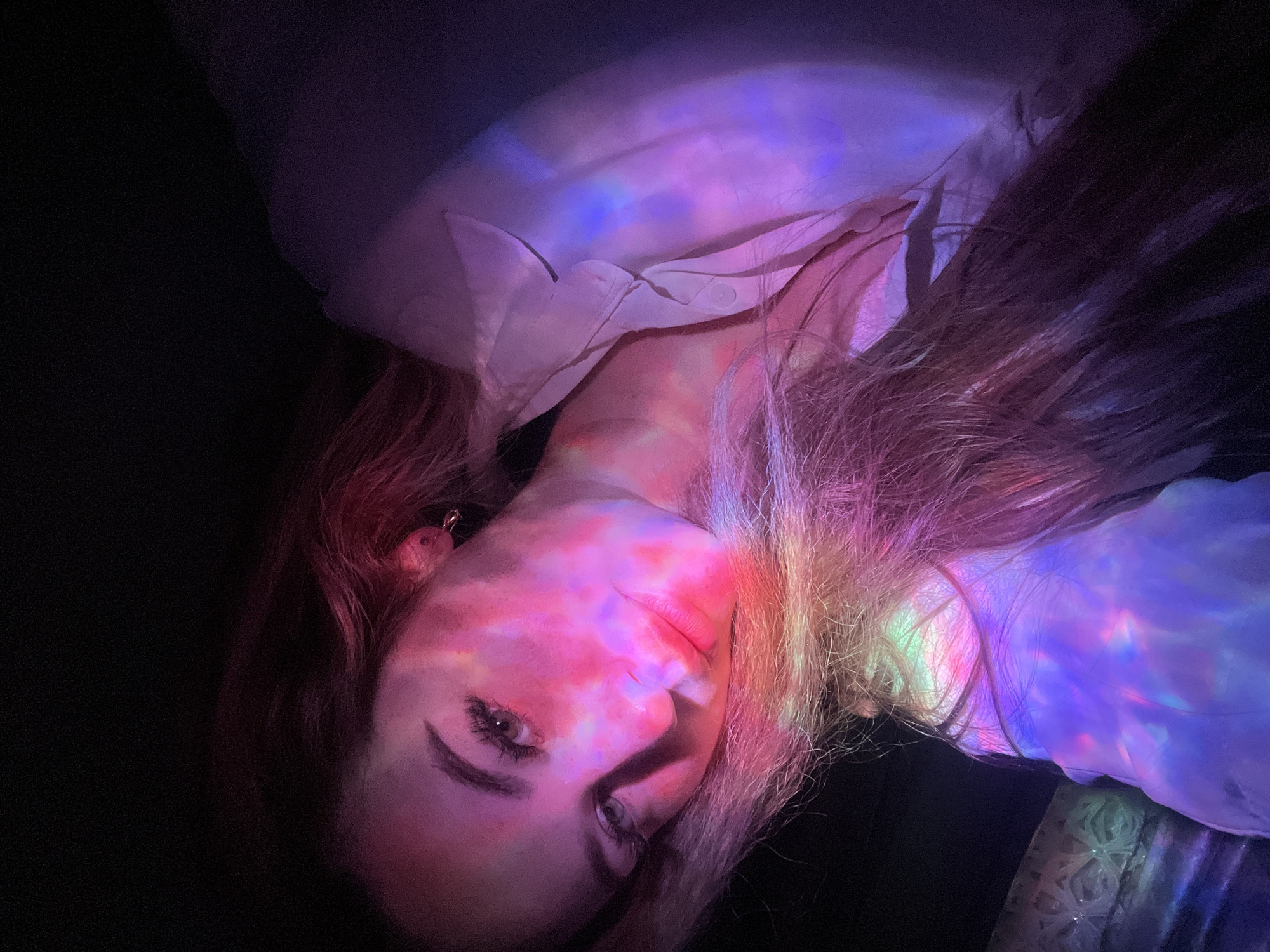 A woman lies in a dark room, possibly indoors, with colorful lights projected onto her face, hair, and clothing. The light patterns include shades of pink, purple, blue, and green. She is wearing a light-colored shirt, possibly white or light pink. The background is dark and obscured. It is impossible to confirm the specific location of Chaikovsky, Russia based on this image alone.