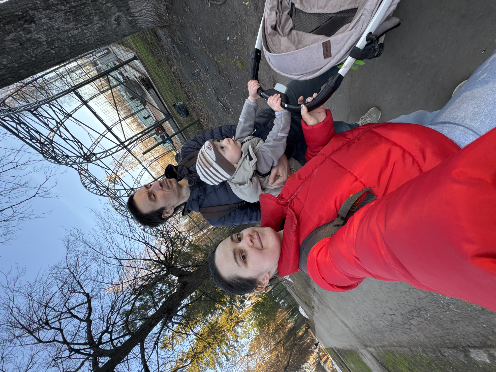The image captures three individuals outdoors, likely a family, during what appears to be a cool season walk in Izhevsk, Russia. In the foreground, a woman wearing a vibrant red puffer jacket and light blue jeans is seen pushing a beige baby stroller. She looks towards the camera with a slight smile. Behind her, a man in a dark jacket is visible, looking upwards or forward. Seated in the stroller is a young child, dressed in a striped beanie and a grey puffer jacket, holding onto the stroller's handle and also gazing upwards with a curious expression. The brand "JOGAREST" is visible on the stroller's fabric.
The setting features a paved path and a backdrop of bare trees, indicating late autumn, winter, or early spring. The sky is clear and bright, suggesting daytime. A significant architectural element dominates the background: a large, intricate metal structure, possibly a roller coaster or a decorative archway, which seems to be the focus of attention for the man and child. A fence is also discernible further back. The scene conveys a moment of family activity and observation in an outdoor environment.