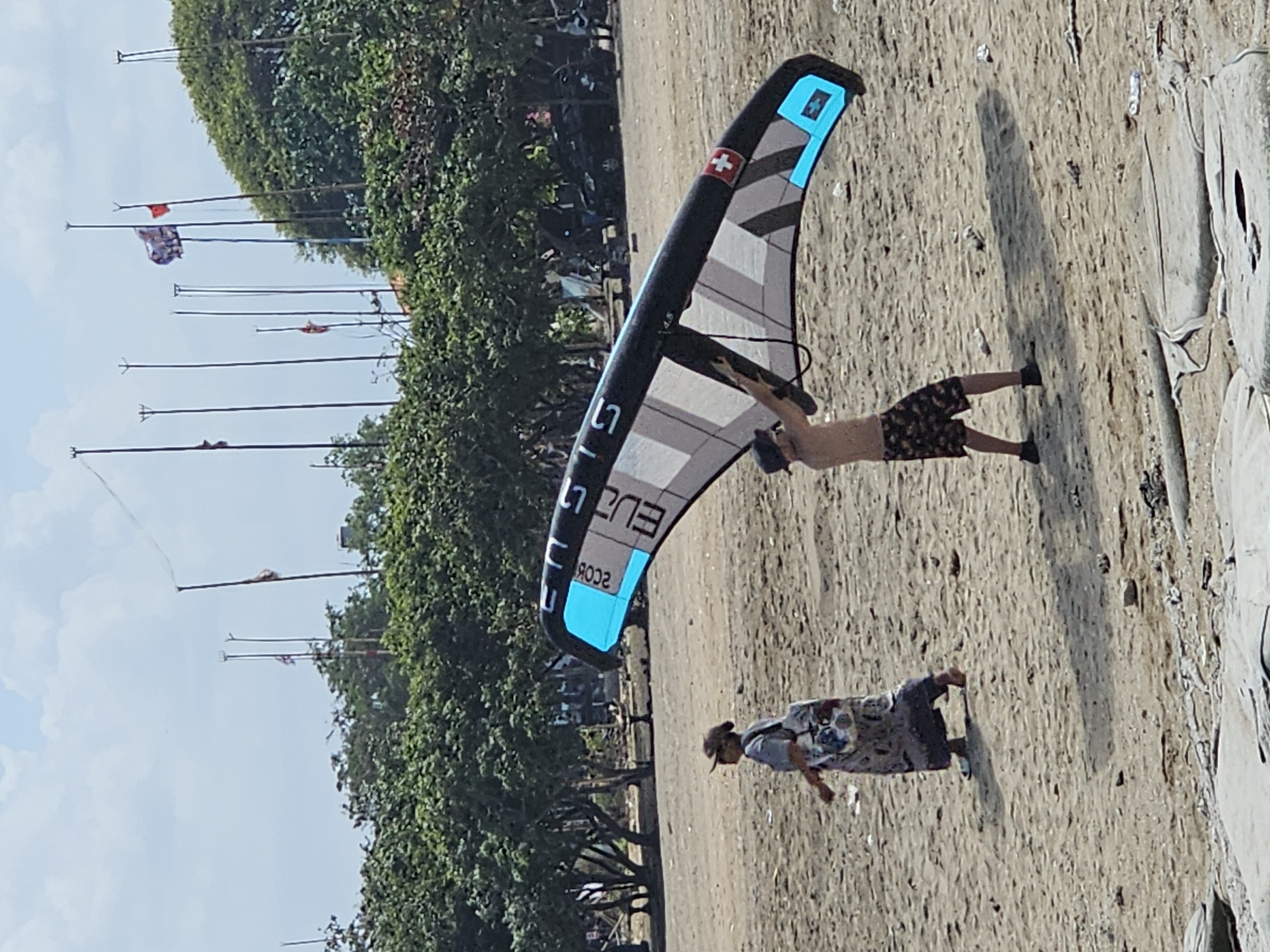 On a sandy beach in Denpasar, Indonesia, two individuals are engaged in activities related to kitesurfing. In the foreground, a shirtless person wearing patterned shorts and a hat is holding up a large kitesurf kite. The kite, predominantly black and white with blue accents and a Swiss flag emblem, is positioned in a way that suggests it's being prepared for use or has just been landed. To the left, a person in a long, patterned dress and a hat walks across the sand, carrying a bag over their shoulder. The background is characterized by lush green trees, a bright sky with scattered clouds, and several flagpoles, some of which display flags. The sandy terrain of the beach is littered with small debris.