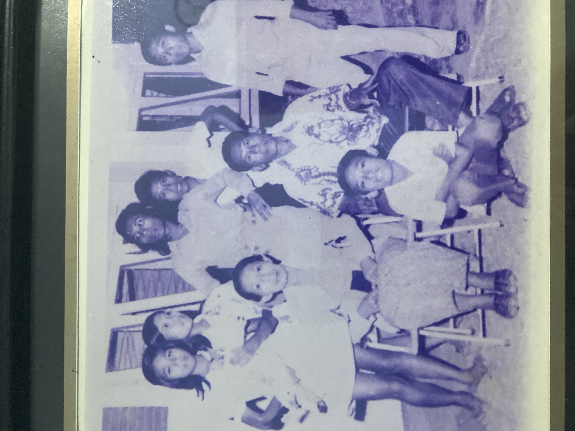 The provided image is a black and white photograph featuring a group of nine individuals, consisting of adults and children, posing outdoors. The setting appears to be in front of a building with visible windows and vertical elements, possibly part of a house or a communal structure.
The individuals are arranged in two rows. In the back row, from left to right, are three young females and three adult males. The two leftmost females are wearing dresses, and the third is partially obscured. The males behind them are dressed in light-colored shirts. The front row features an adult female seated in a chair on the left, an adult male seated in a chair in the center wearing a patterned shirt, and a young male crouching in front of him. To the right of the seated adult male, another adult male stands.
The environment is somewhat sparse, with a concrete or dirt ground. The lighting suggests it is daytime. There are no specific landmarks or visual cues that definitively place this photograph in Kediri City, Indonesia. The overall impression is a casual, posed group photograph, likely taken in a residential or community setting.