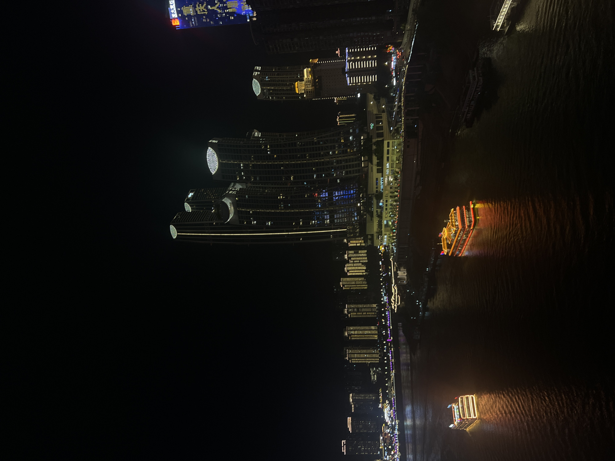 The image captures a nighttime cityscape along a wide river, likely in Yuzhong District, China. The foreground features two illuminated tour boats navigating the dark water, their lights reflecting on the rippling surface. The boat on the left is rectangular with multiple decks and is outlined in warm white lights. The boat on the right is longer and more elaborate, with red and orange lights illuminating its stacked decks and roof. Further in the distance, the riverbank is lined with a dense cluster of illuminated buildings. Among these are several modern skyscrapers, some with distinctive curved designs and lit tops. One tall building on the right is adorned with bright blue and yellow vertical lights, displaying Chinese characters. The overall scene is one of urban vibrancy and nighttime activity on the water and along the riverfront.