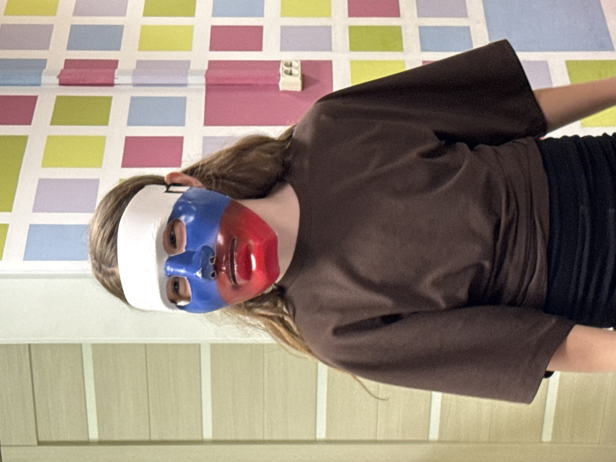 A person wearing a white, blue, and red mask is the central figure in this photo. The mask covers the individual's face, with blue coloring the left side, red on the right, and a white strip across the forehead. The person is wearing a brown, loose-fitting top and black pants.
The background consists of two distinct sections. The wall behind the person is decorated with a multicolored geometric pattern of rectangles in shades of pink, yellow, blue, and lavender, and an electrical outlet is visible on the wall. The lower portion of the background appears to be a wall with vertical panels in a light tan color.
The image's location is presumed to be in Chaikovsky, Russia, based on the tri-color mask that replicates the Russian flag.