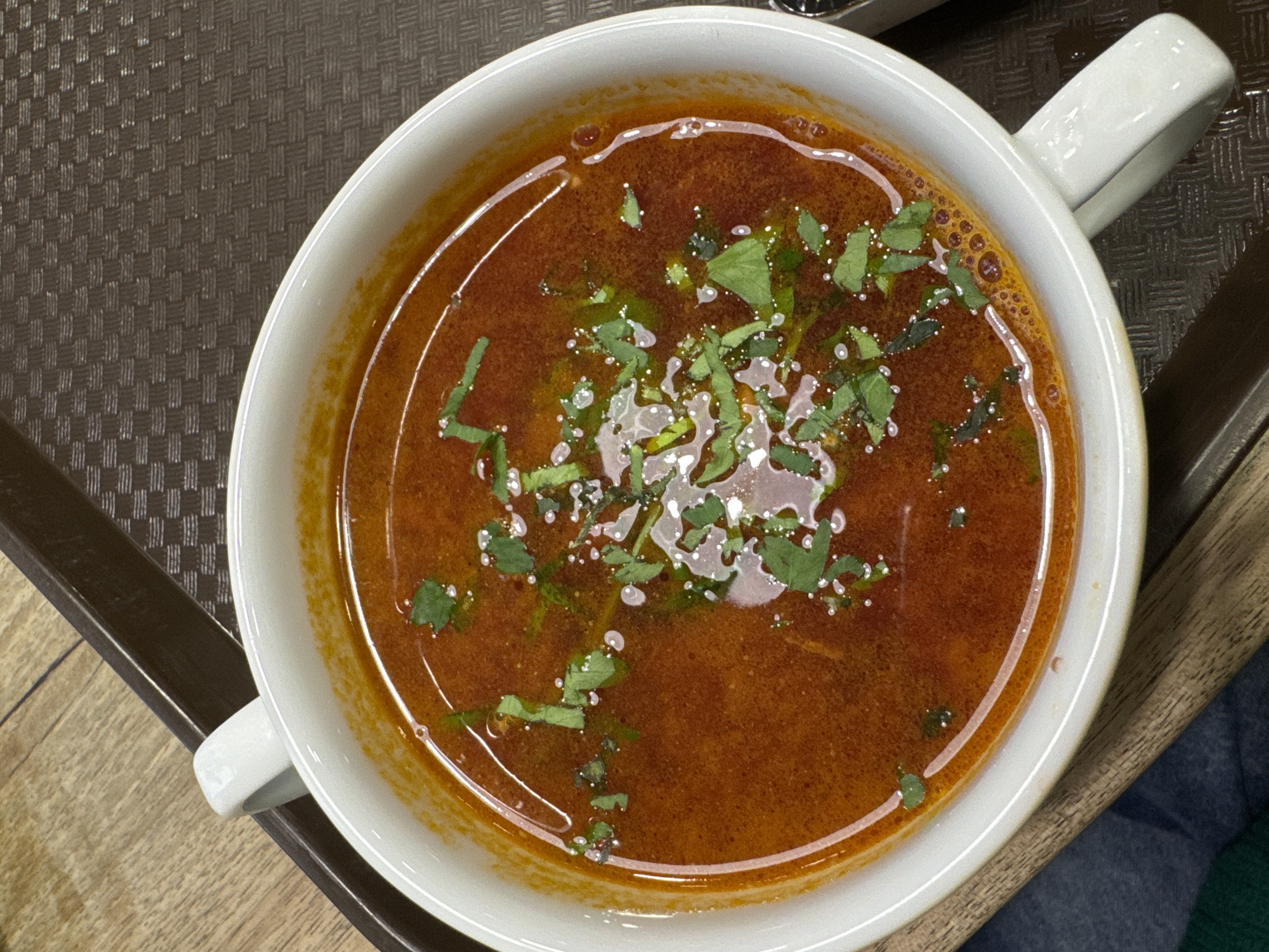 A top-down, close-up shot depicts a white mug filled with a dark red, broth-like soup garnished with scattered green herbs. The mug has a wide, rounded opening and a handle extending to the right. The soup appears slightly oily with small bubbles and scattered herbs. The mug sits on a surface with a dark brown, woven texture. Patches of wood-colored surface and parts of a dark brown frame edge appear around the mug. The location of this scene is not identifiable.
