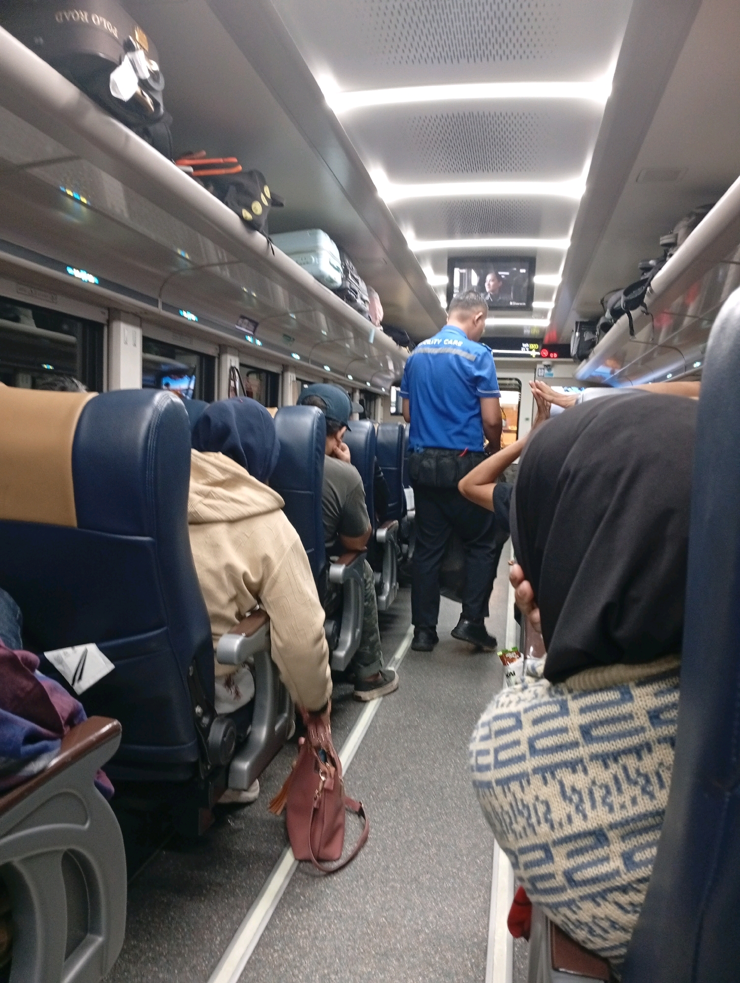 The image depicts the interior of a passenger train, likely a commuter or long-distance service, with passengers seated and luggage stored overhead. A man in a blue uniform with "SECURITY CARE" visible on the back is standing in the aisle, facing away from the camera. He is wearing a dark belt with pouches. To his left, a woman in a dark hijab and a patterned sweater is seated, her back to the camera. In the foreground, the back of a blue train seat is visible. Other passengers are seated in the background, some with their heads covered or turned away. Luggage, including suitcases and bags, is stowed on overhead racks. The train carriage appears to be well-lit with overhead fluorescent lighting. The floor is a speckled grey material. The overall impression is one of a typical journey on public transport. The specific location in Kubangkarang, Indonesia, cannot be determined from the visual information in the image alone; this detail would require external context or identification of specific train line markings not present.
