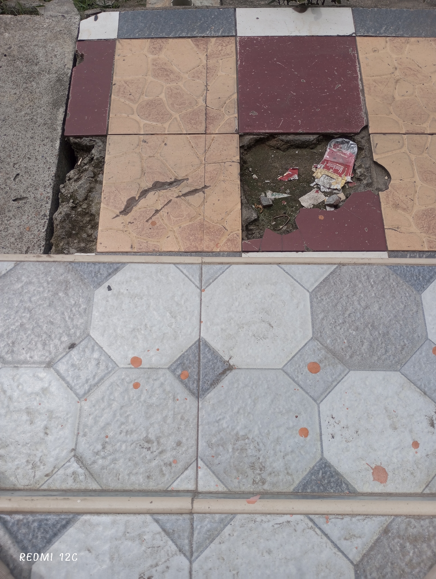 The image depicts damaged tiled flooring in Lhokseumawe, Indonesia. The upper section shows a pattern of alternating red and tan square tiles, with several tiles missing or broken, revealing a cavity filled with debris and a discarded Marlboro cigarette packet. Adjacent to this is a concrete surface. The lower section exhibits a different tile pattern of light grey octagons with dark grey squares at the corners, and several reddish-brown stains are visible on the surface. A tan border tile separates the two tiled sections. The lower left corner of the image includes the text "REDMI 12C".