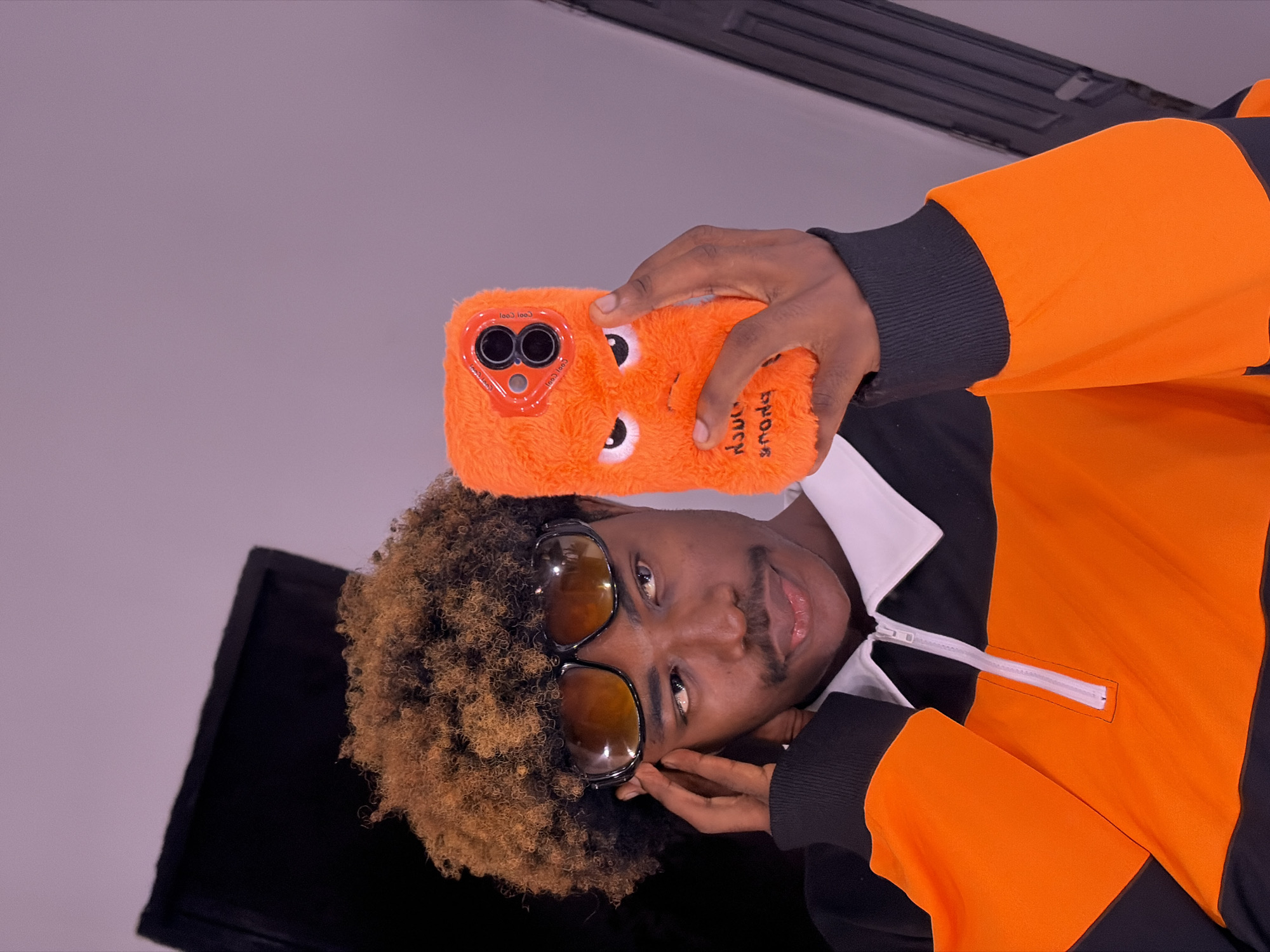 A man with light brown afro-style hair, wearing sunglasses and a black and orange shirt with a white collar, holds an orange, furry phone case with cartoon eyes in front of his face. The background is a plain, light gray wall with a dark vent near the top and a black, reflective surface at the bottom left. The location is unspecified, but the city of Igboefon, Nigeria, is associated with this scene. The man is looking directly at the camera, holding the phone as if taking a selfie.